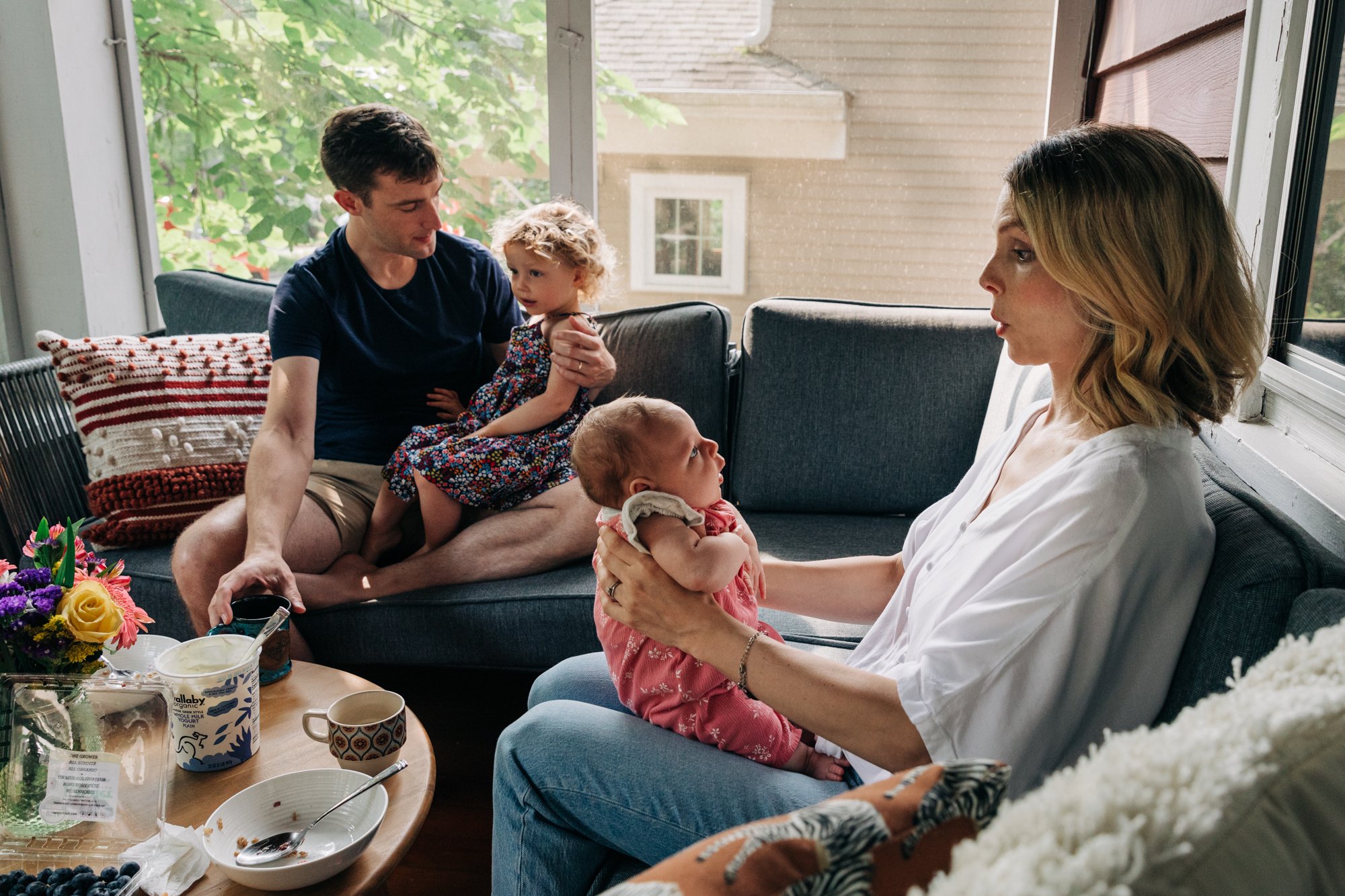 Family sitting on a couch in a sunlit room, with a man holding a young girl and a woman holding a baby girl, near a window with trees outside.