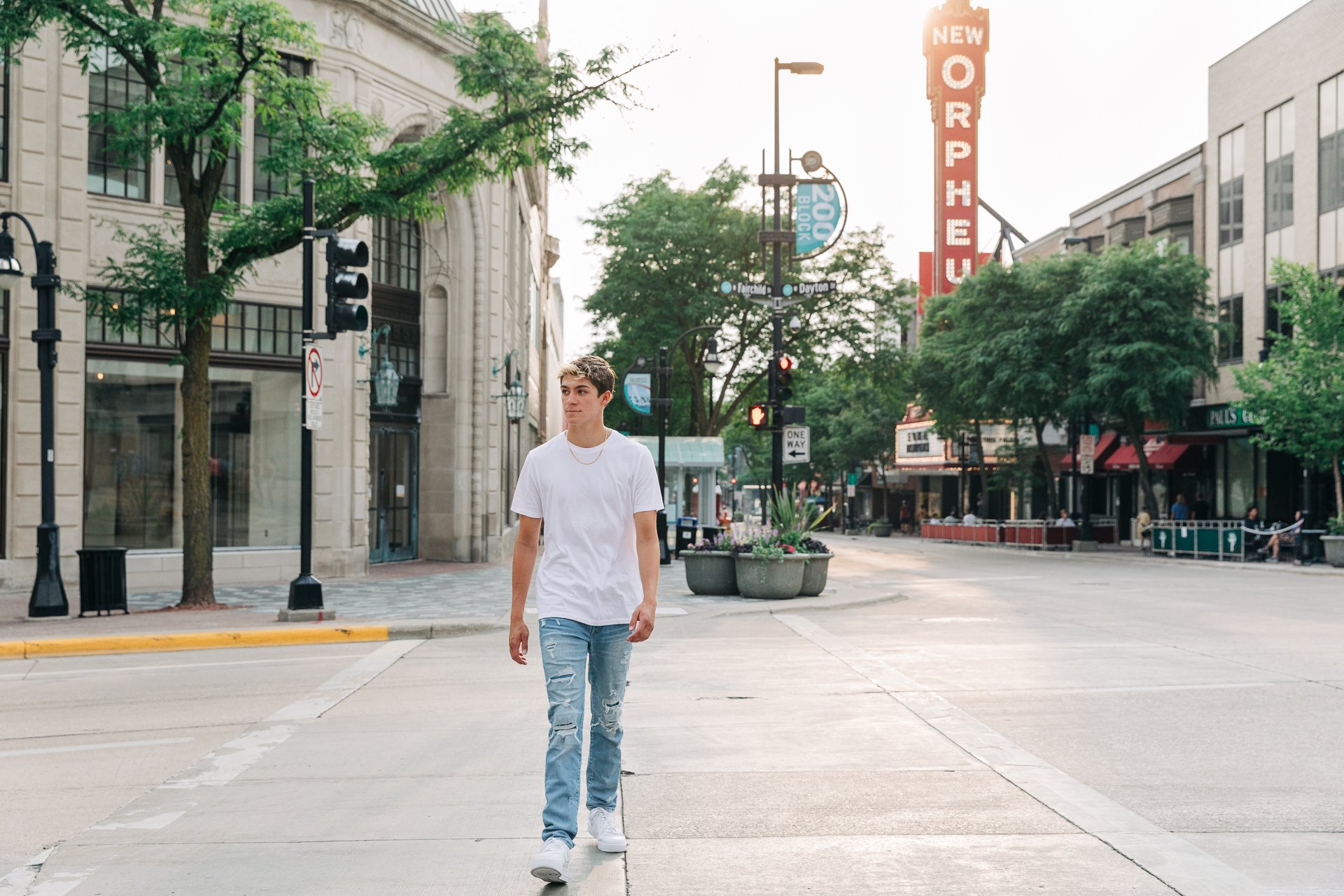 A young man walking on a city sidewalk in the early evening with empty streets, trees, and buildings in the background.