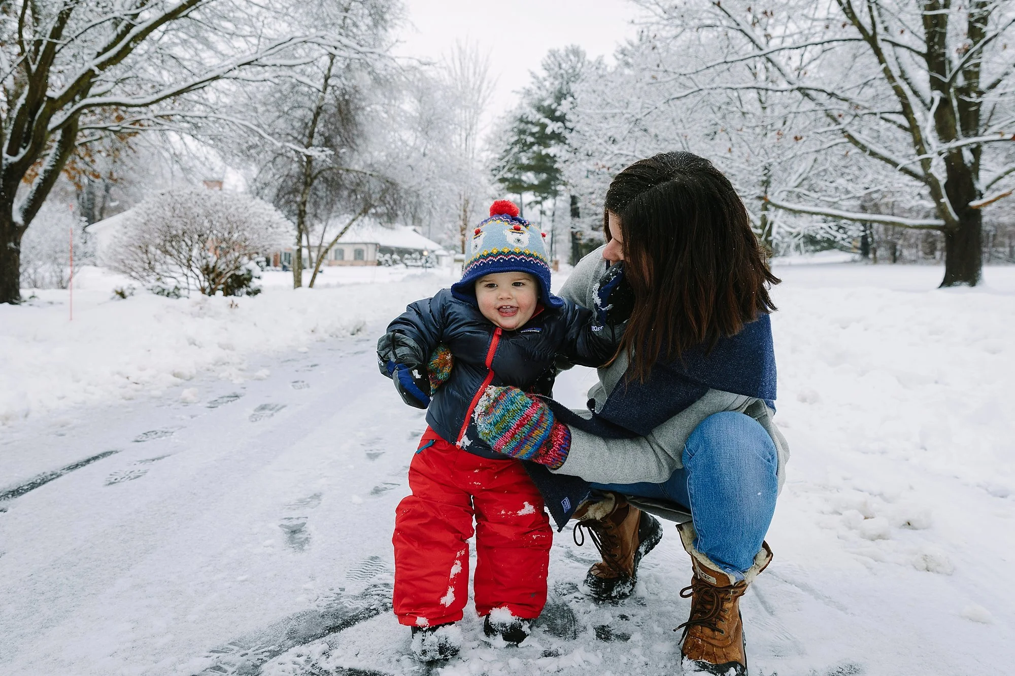 A woman and a young child playing in the snow on a winter day, with trees and houses in the background.