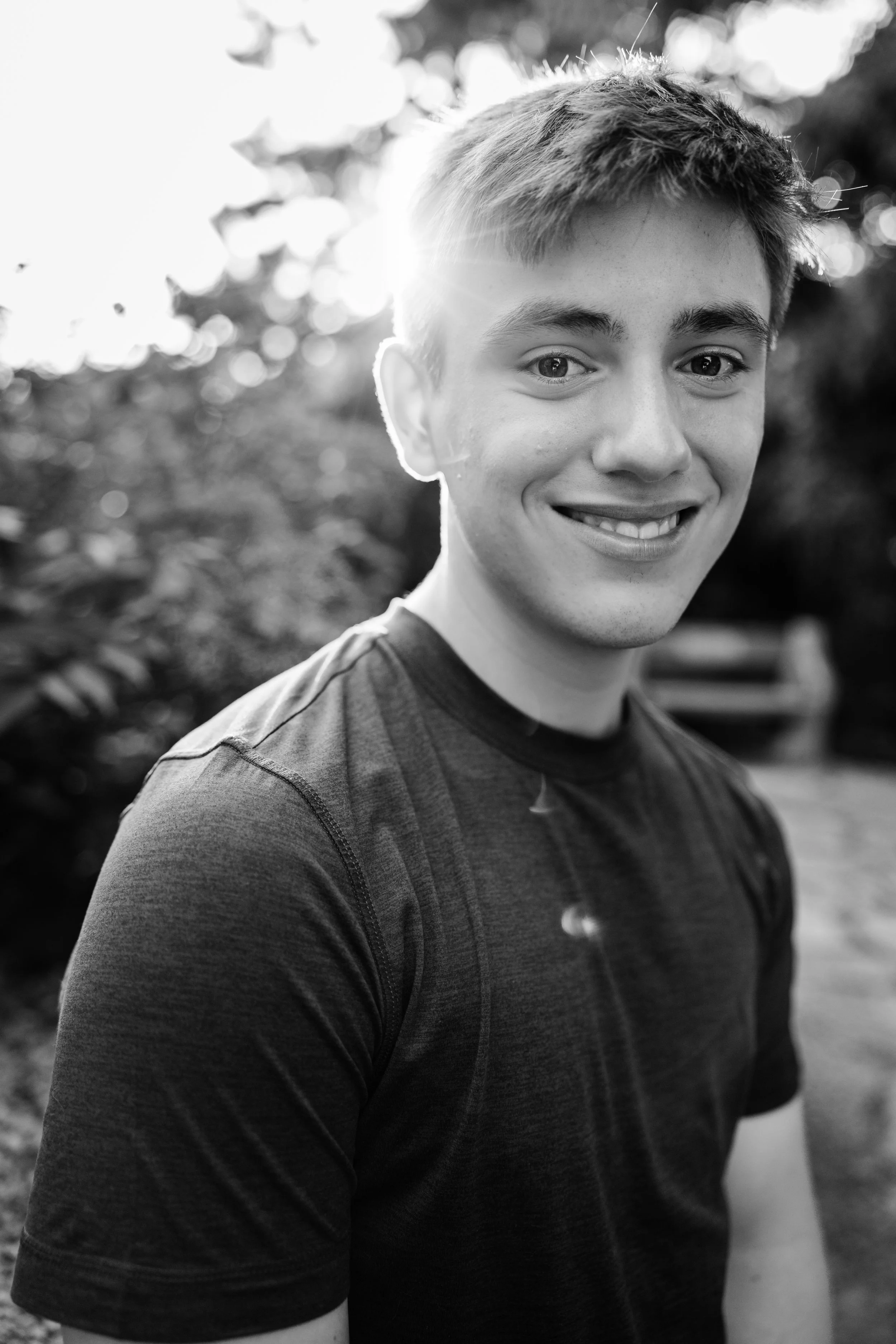 Black and white photo of a young man smiling outdoors, wearing a dark athletic shirt, with sunlight behind him and blurred trees in the background.
