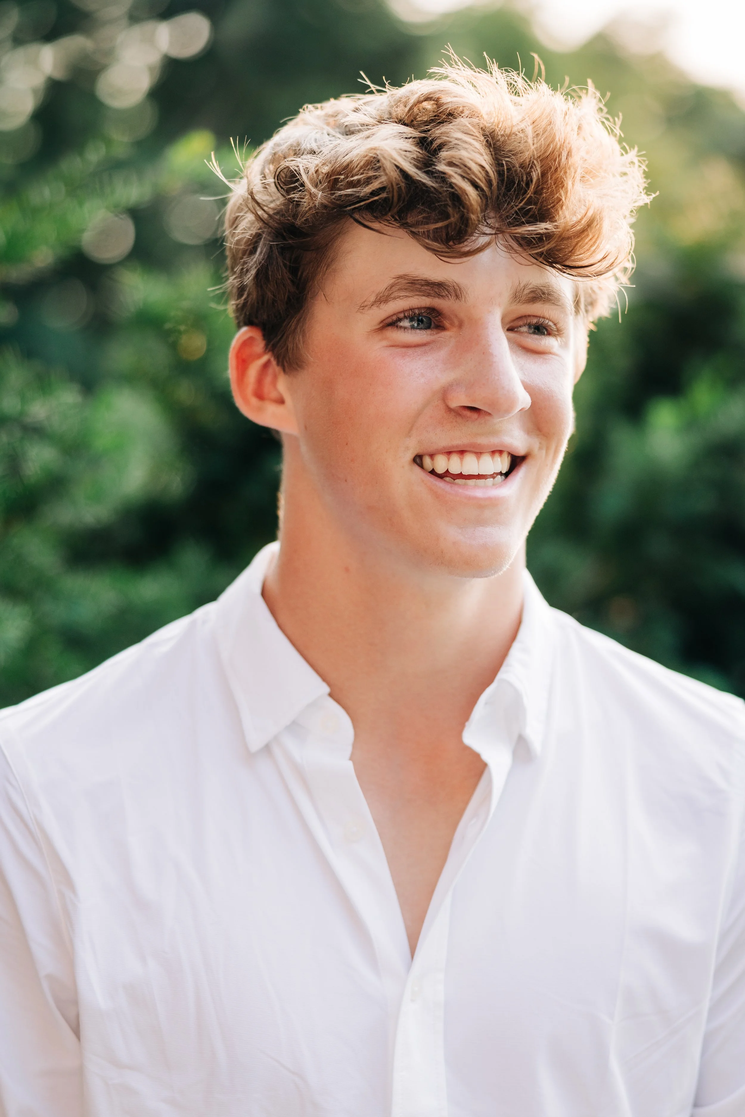 A smiling young man with wavy brown hair and blue eyes, wearing a white button-up shirt, standing outdoors with greenery in the background.