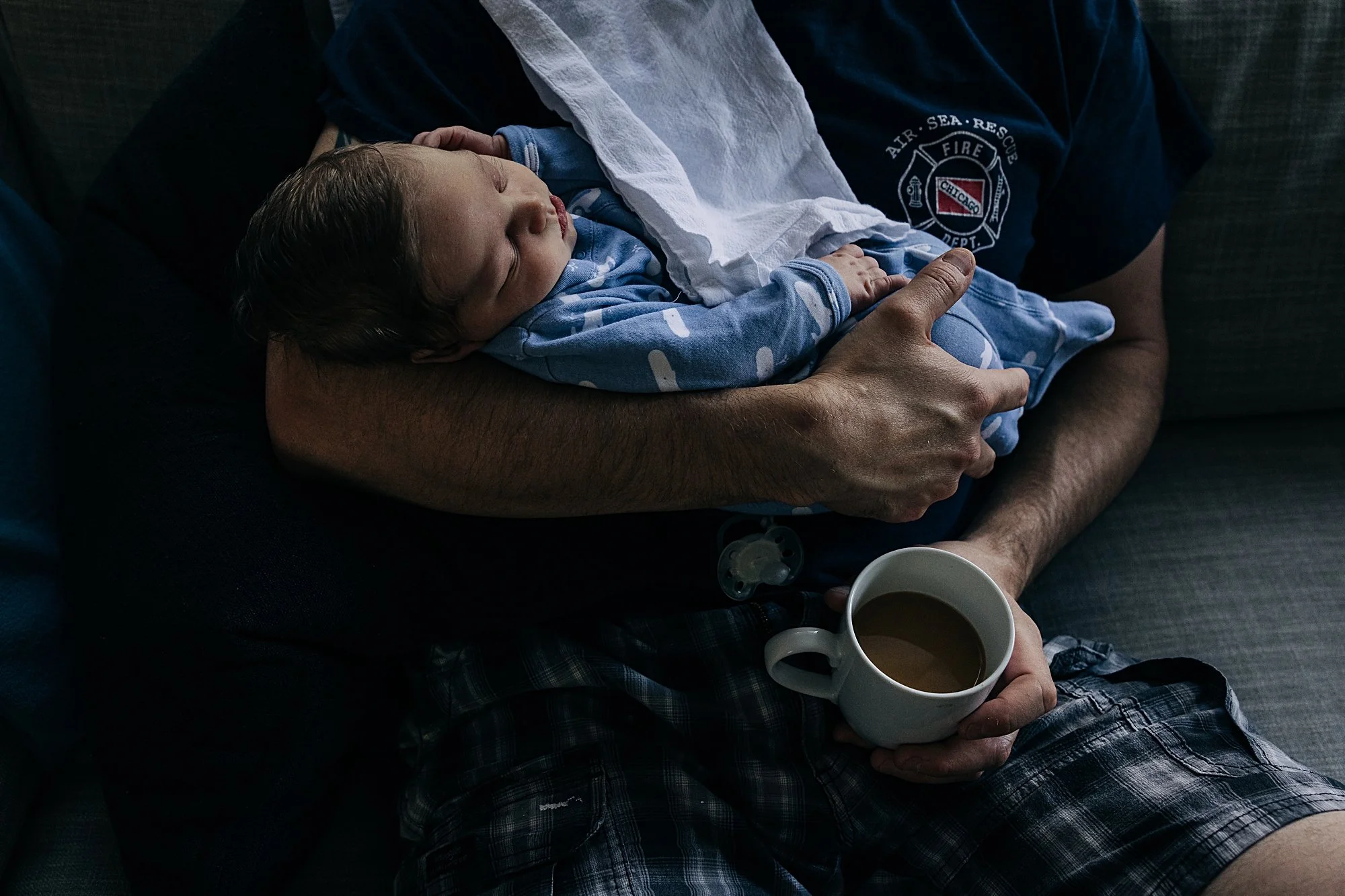 A man holding a sleeping baby on his chest, sitting on a couch with a mug of coffee in his hand.