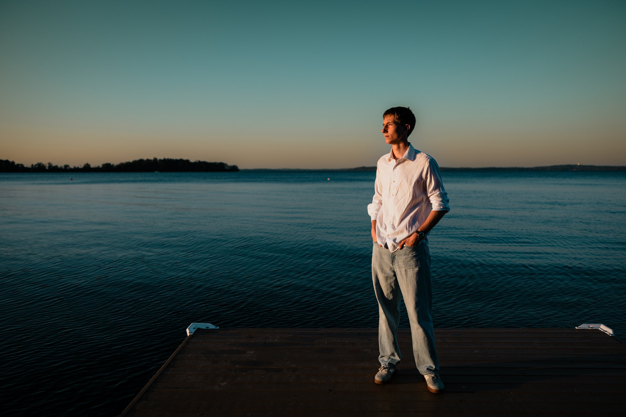 A young man in a white shirt and light jeans stands on a dock by a calm lake during sunset, with his hands in his pockets, looking thoughtfully at the water.