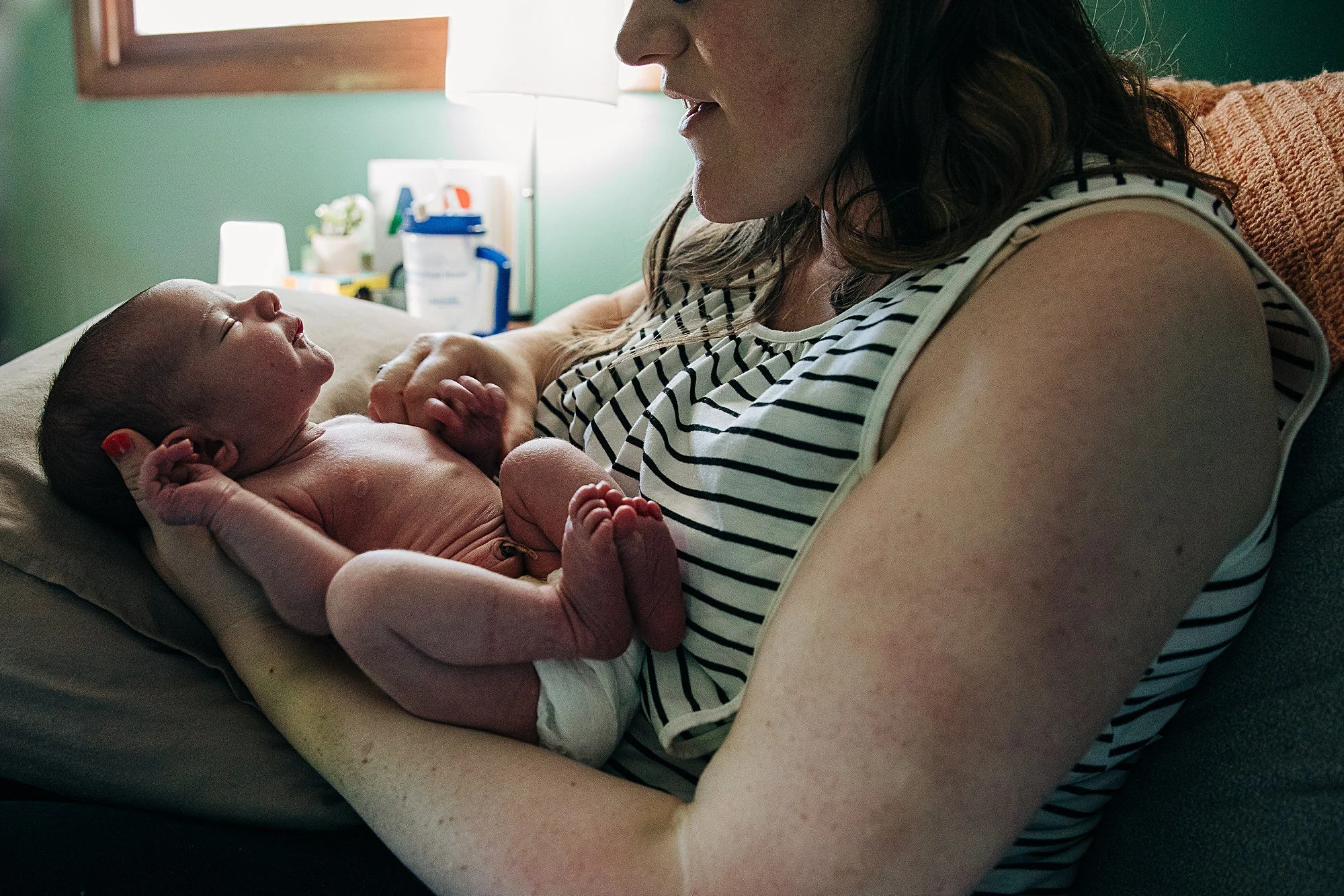 A woman with dark hair and freckles wearing a sleeveless striped shirt holds a newborn baby in her arms, lying on a hospital bed, with the baby smiling and closed eyes. The background has a green wall, a bedside lamp, and medical supplies.