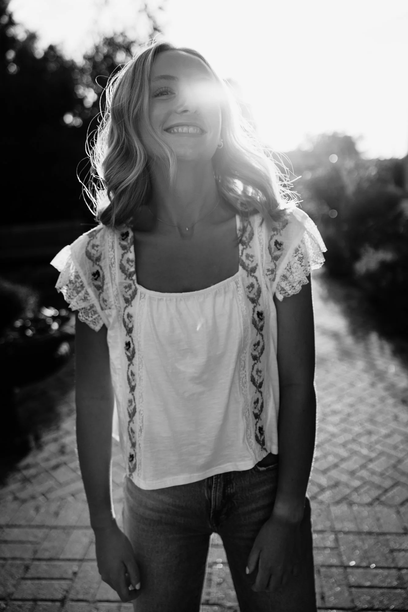 Black and white photo of a smiling woman standing outdoors in sunlight, wearing a white embroidered top and jeans, with trees and a paved path in the background.