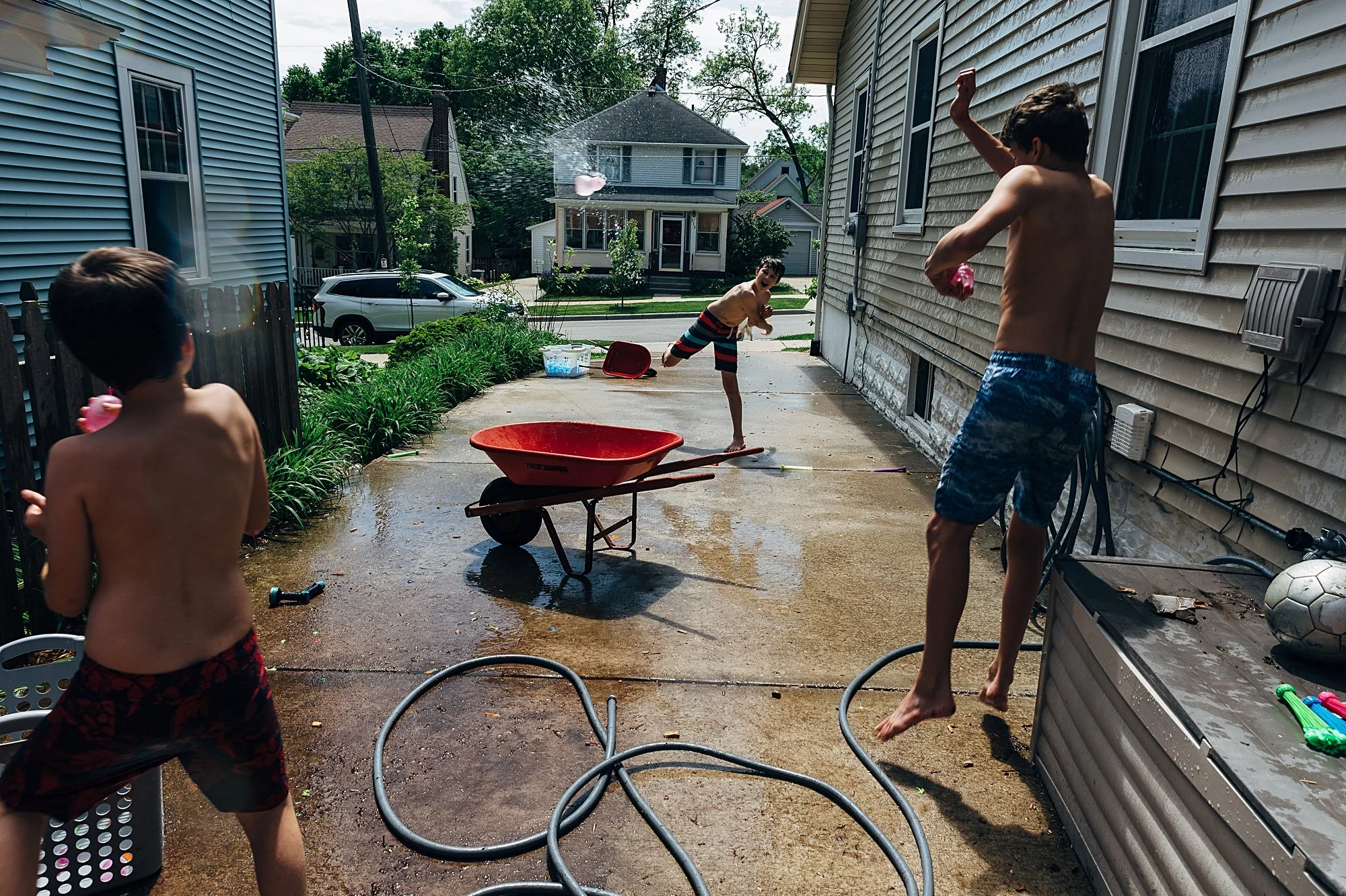 Three boys playing with water guns and a water balloon on a backyard concrete patio, with a wheelbarrow and garden hose present, during daytime on a sunny day with trees and houses in the background.