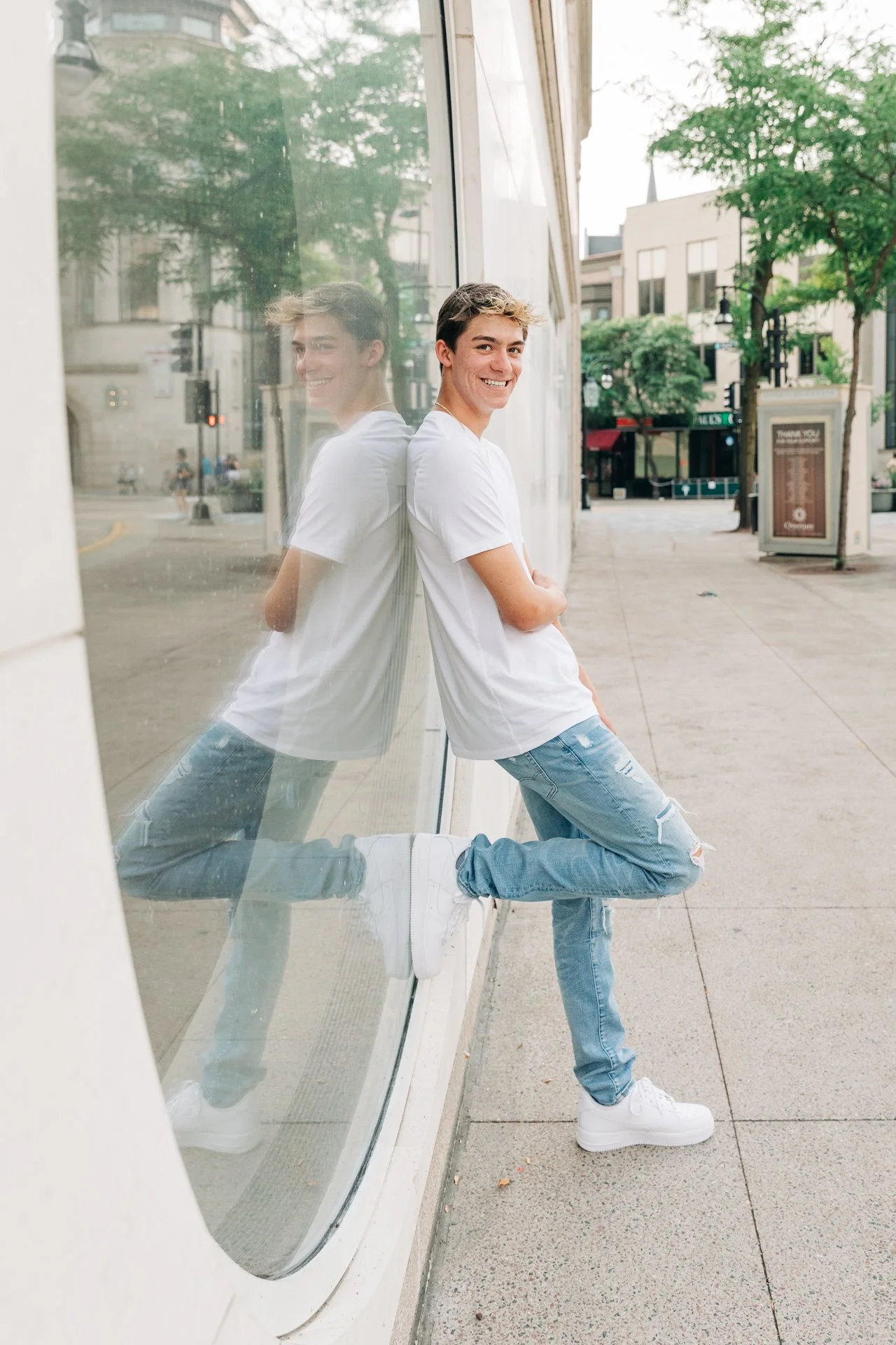 Young man with short brown hair smiling, leaning against a glass window on a city sidewalk, with his reflection visible in the glass, wearing a white t-shirt, ripped jeans, and white sneakers.