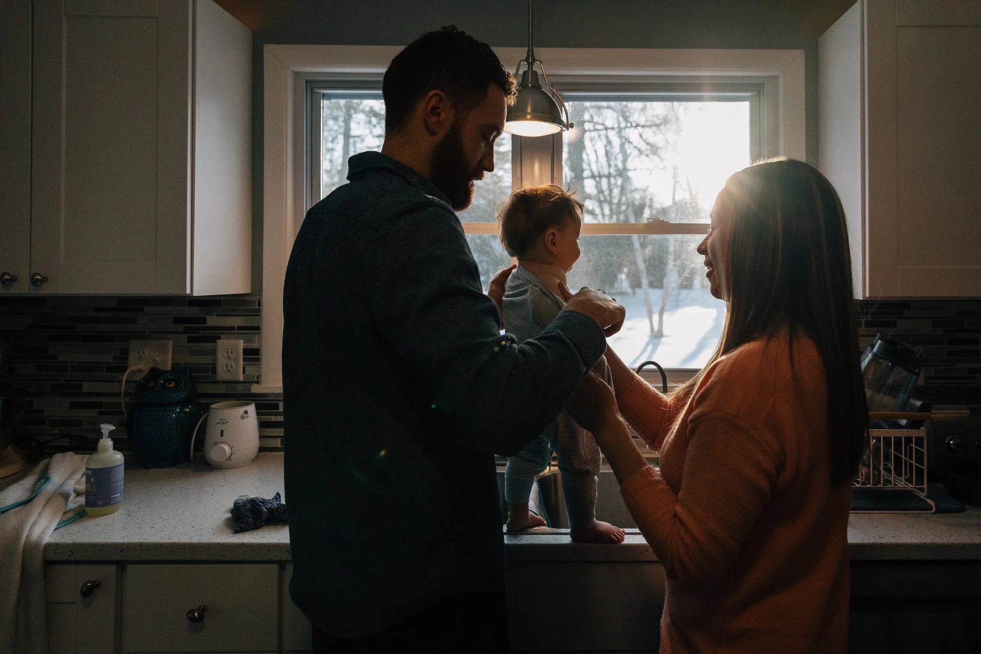 A family of three in a kitchen, with a man and woman holding a baby between them, backlit by a window showing a snowy outdoor scene.
