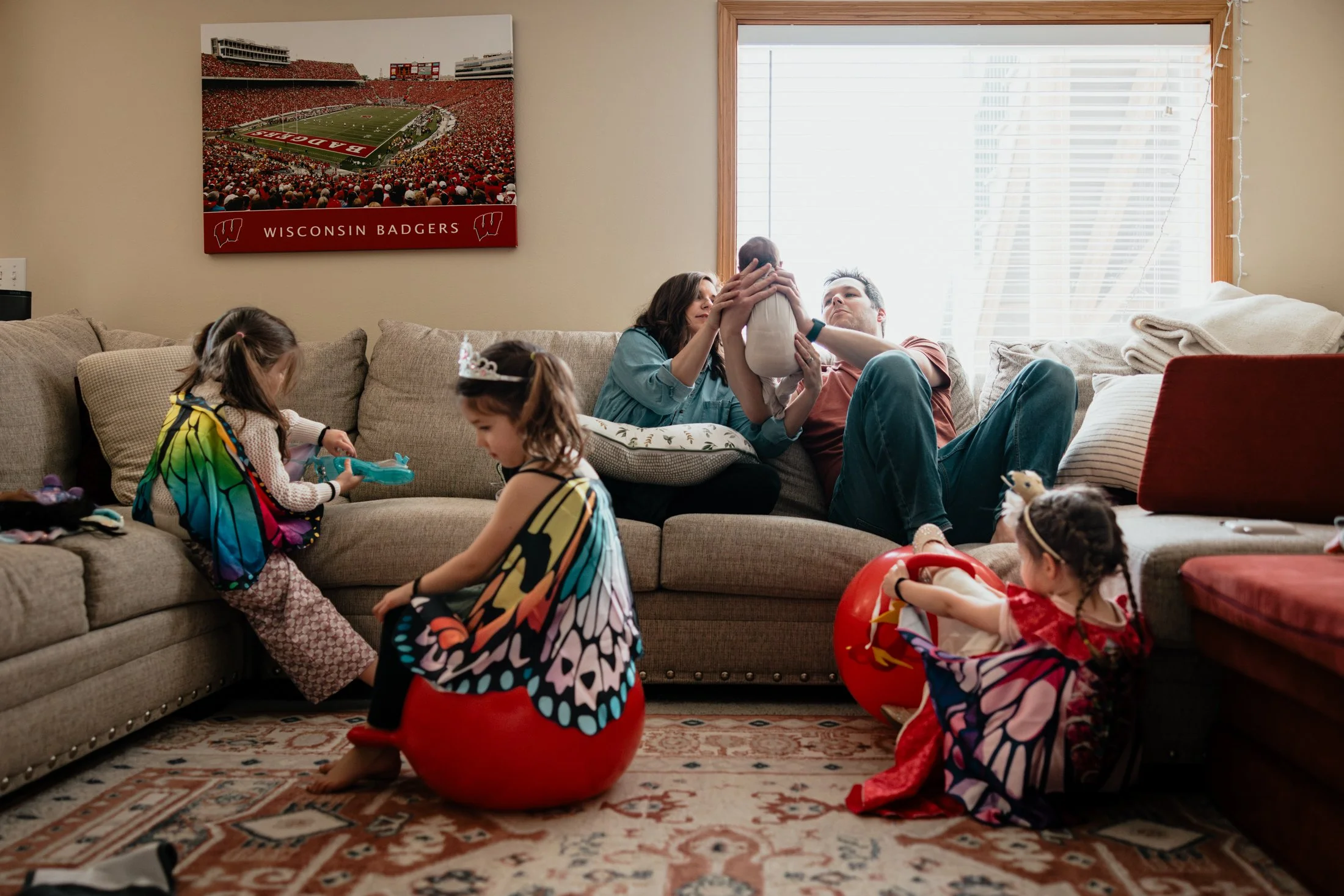 A family with young children in costumes and fairy wings playing in a living room, with two adults sitting on a couch. The children are sitting on plush red balls. One child is on the floor with a doll, and another child is holding a toy. The room ha