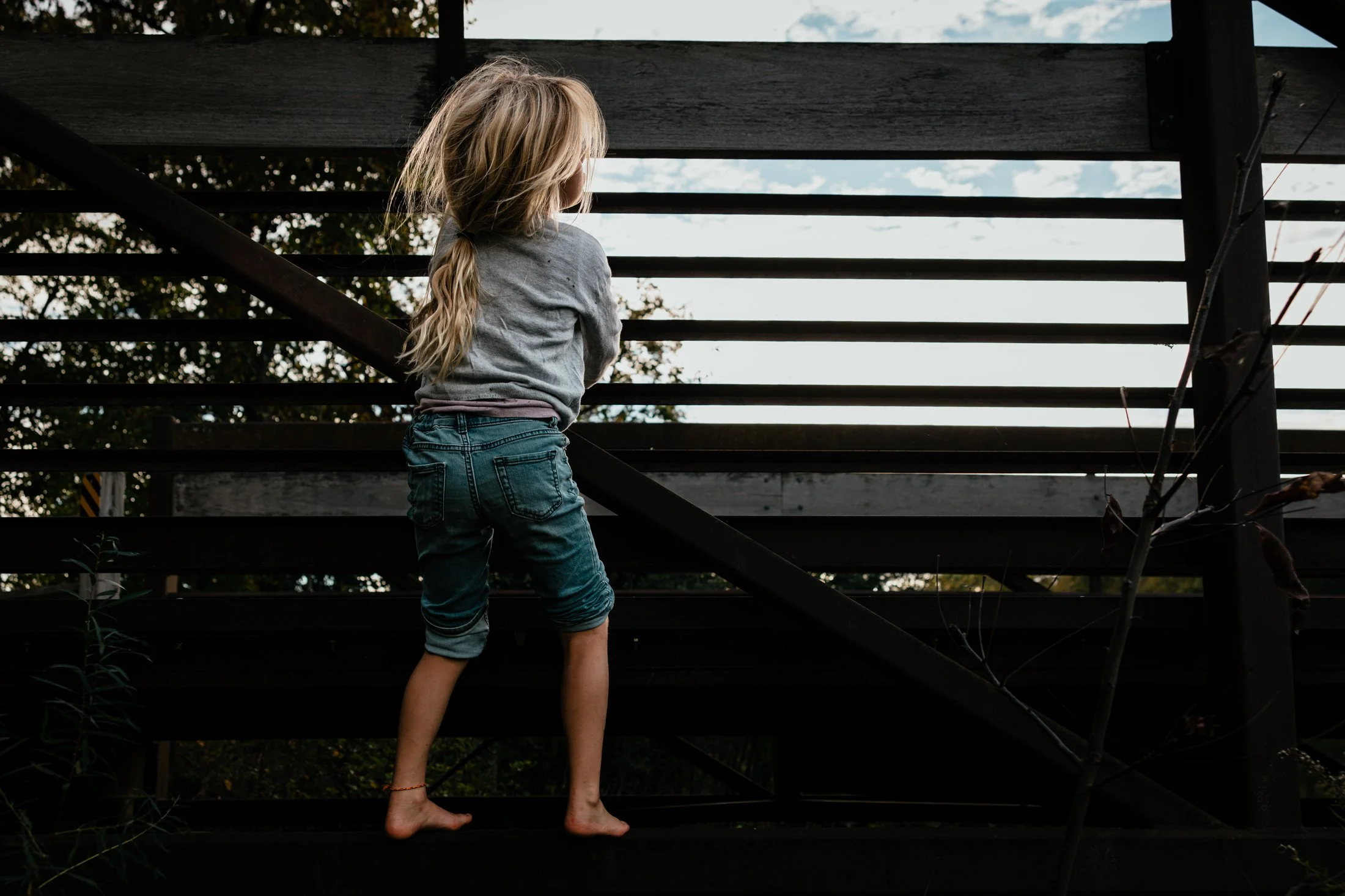A young girl with long blond hair, wearing a gray sweatshirt and rolled-up denim jeans, stands barefoot on a dark outdoor staircase and looks away while holding onto the railing.