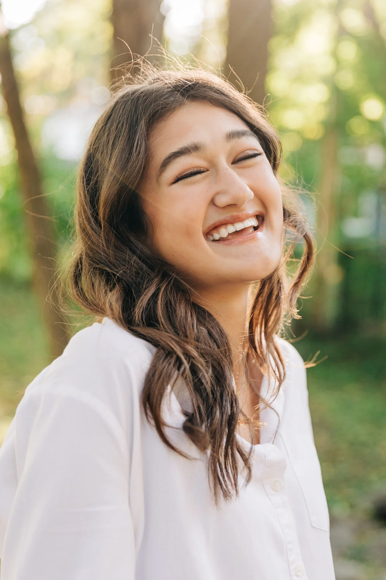 A young woman with wavy brown hair smiling outdoors in a park with green trees and soft sunlight in the background.