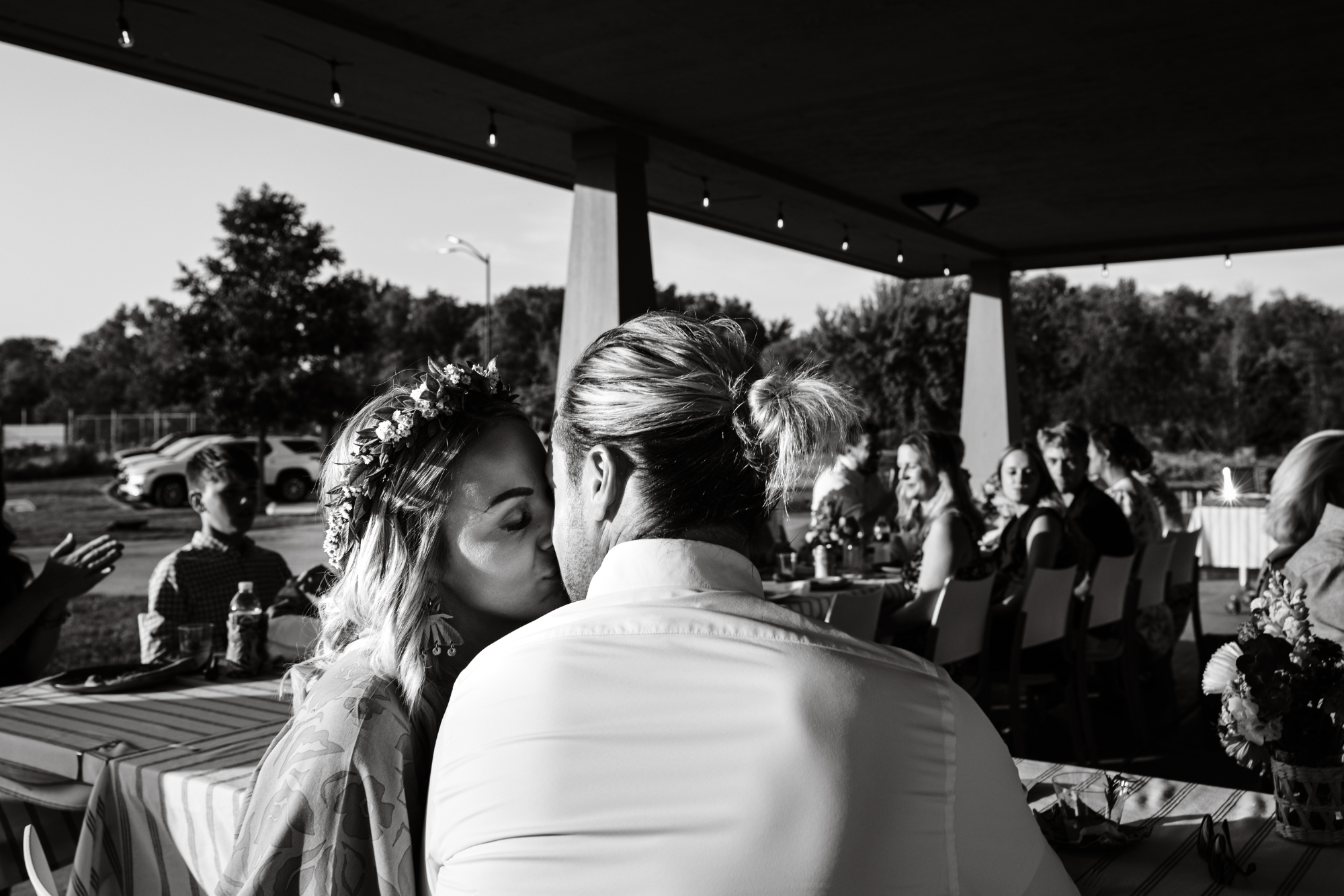 Black and white photo of a wedding reception outdoor under a pavilion, with a couple sharing a kiss at the table in the foreground, and guests sitting and clapping in the background.