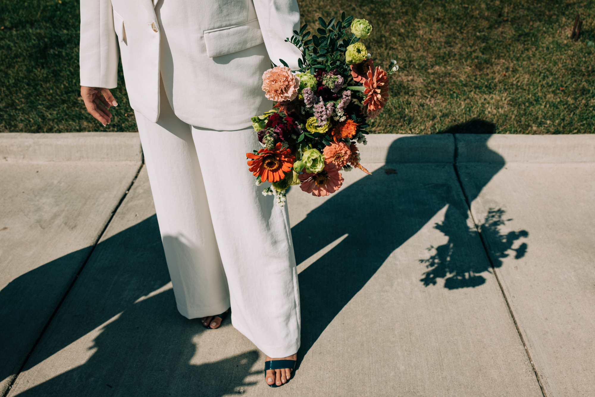 Person in white suit holding a colorful bouquet of flowers on a sidewalk, casting a shadow.