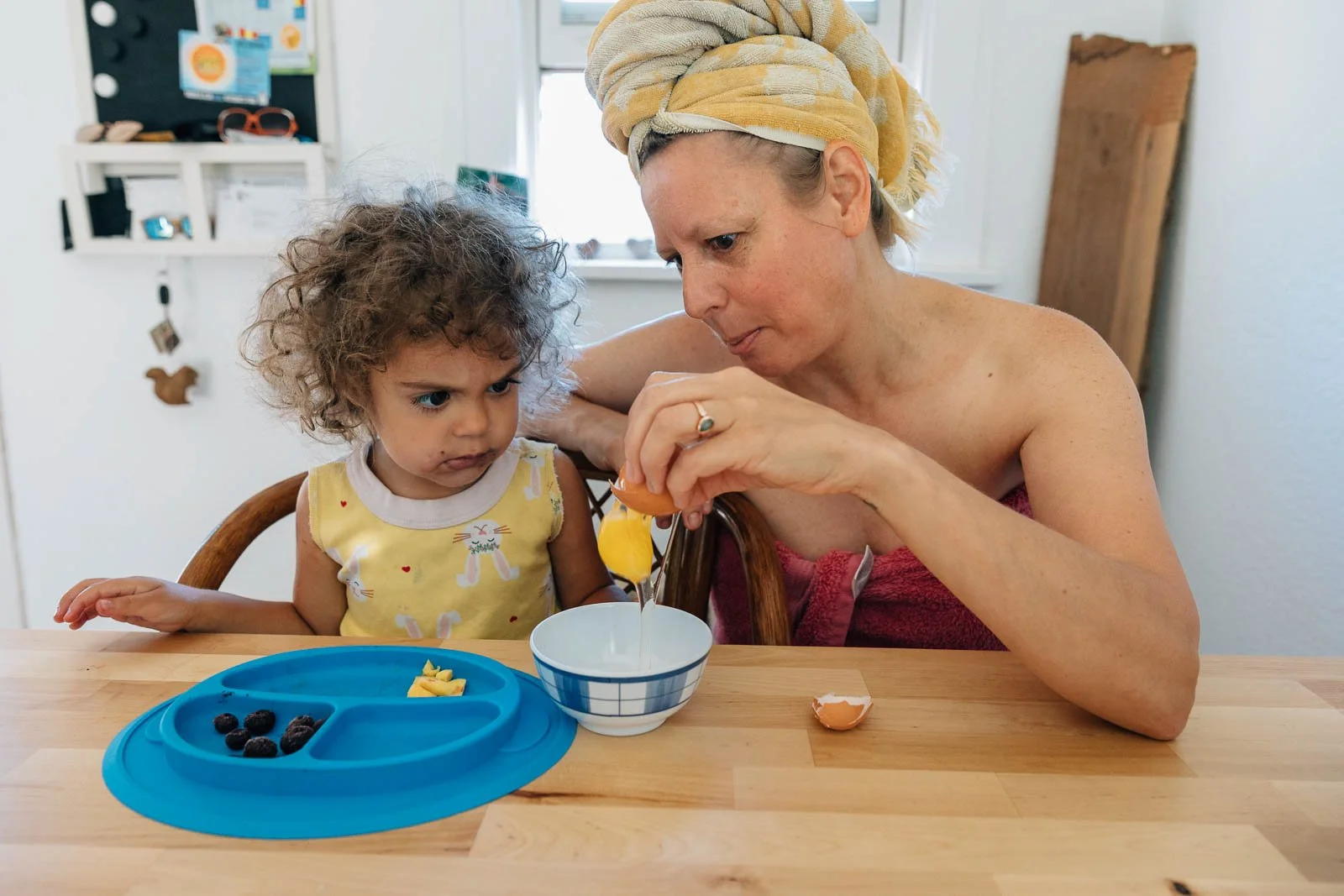 A woman with a towel wrapped around her head and a young girl with curly hair sitting at a wooden table. The woman is cracking an egg into a bowl while the girl looks on with a skeptical expression.