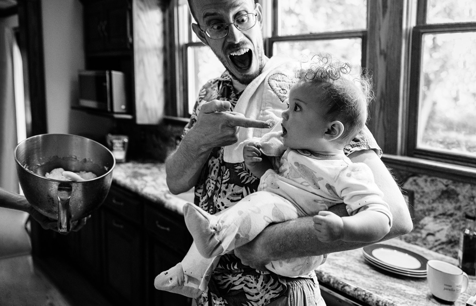 A man with glasses, wearing a patterned shirt, is holding a young child in his arms. The man is excitedly pointing with his finger, and the child, wearing pajamas with dinosaur patterns, is looking at him with a surprised expression. They are in a ki
