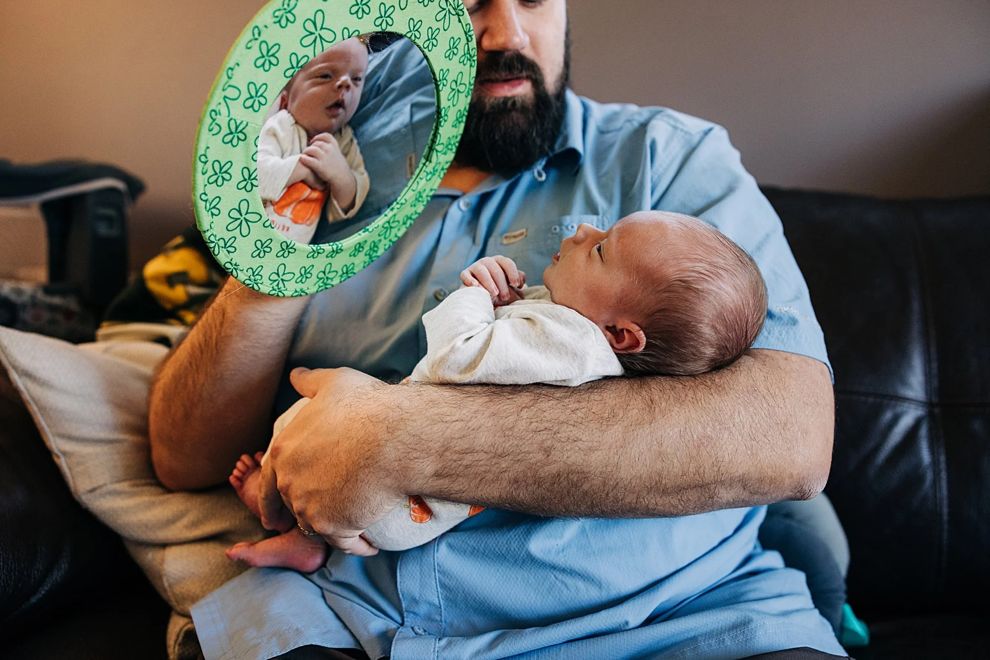 A man with a beard holding a baby in his arms. The man is looking at a small mirror with a green floral frame, reflecting an image of a baby. The baby in his arms is lying on its side, looking up, wearing a white onesie.