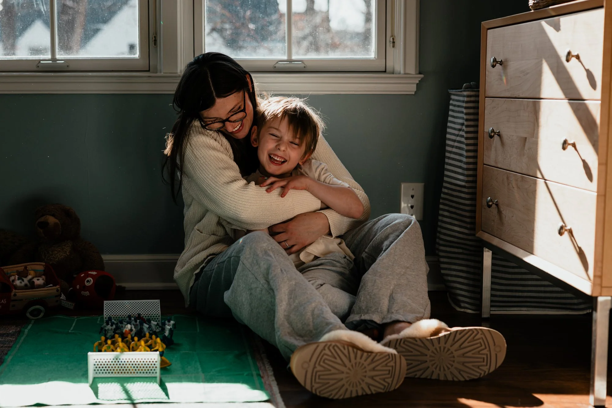 A woman and a young boy are sitting on the floor, laughing and hugging each other, visible next to a window with sunlight coming in. There are toys on a small green mat in front of them and stuffed animals near them in the corner.