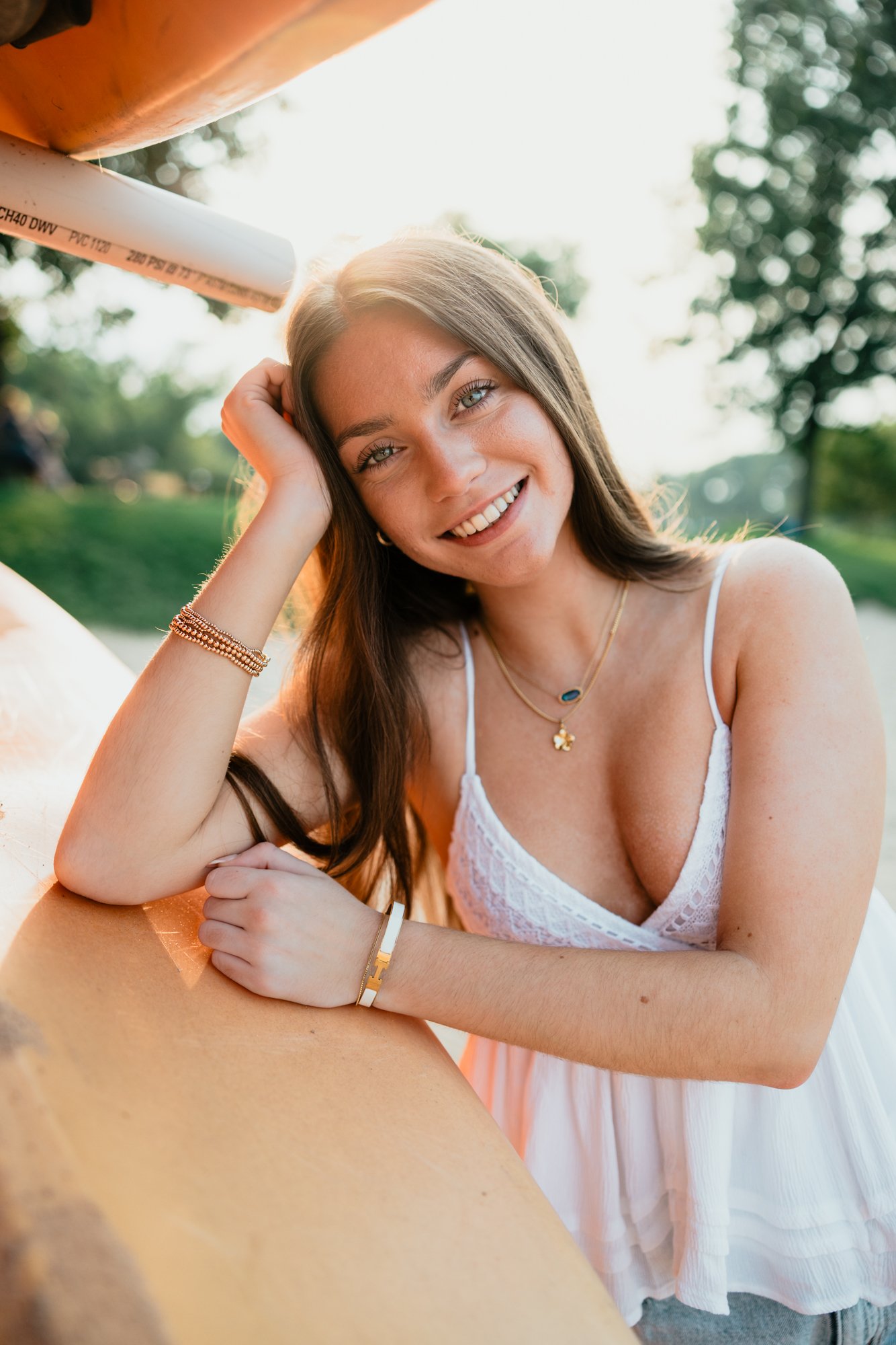 A young woman with long brown hair and blue eyes smiling, resting her head on her left hand, outdoors during sunset, wearing a white lace dress and jewelry, with a blurred green park in the background.