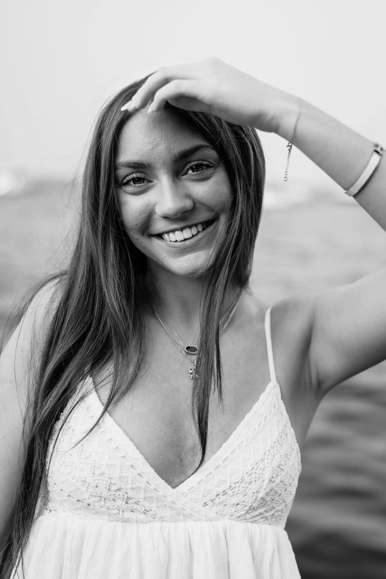 A smiling young woman with long hair, wearing a tank top with lace at the neckline, standing outdoors near water.