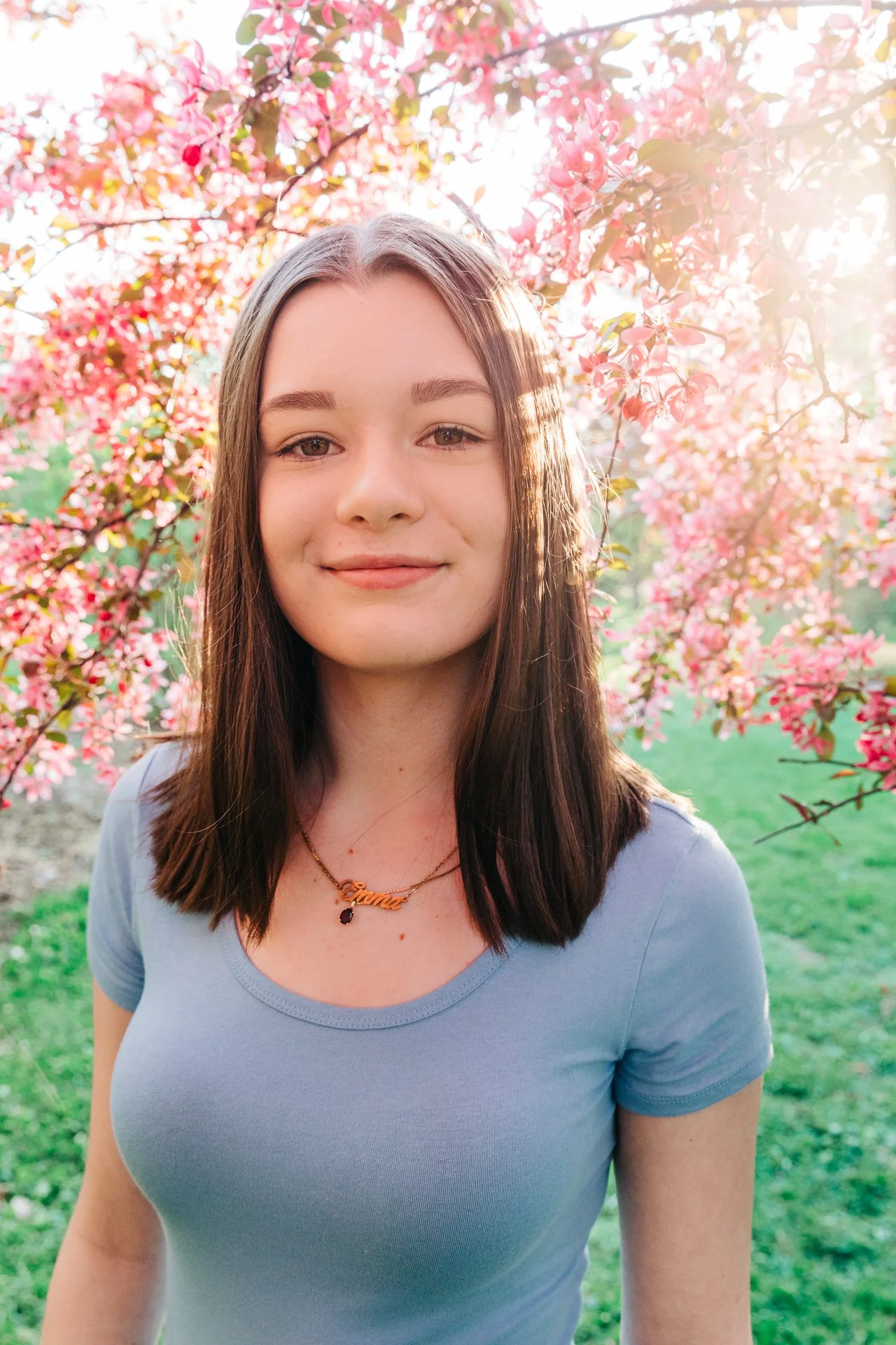 Young woman with brown hair wearing a light blue shirt, standing outdoors in a garden with pink flowering trees, sunlight shining through the branches.