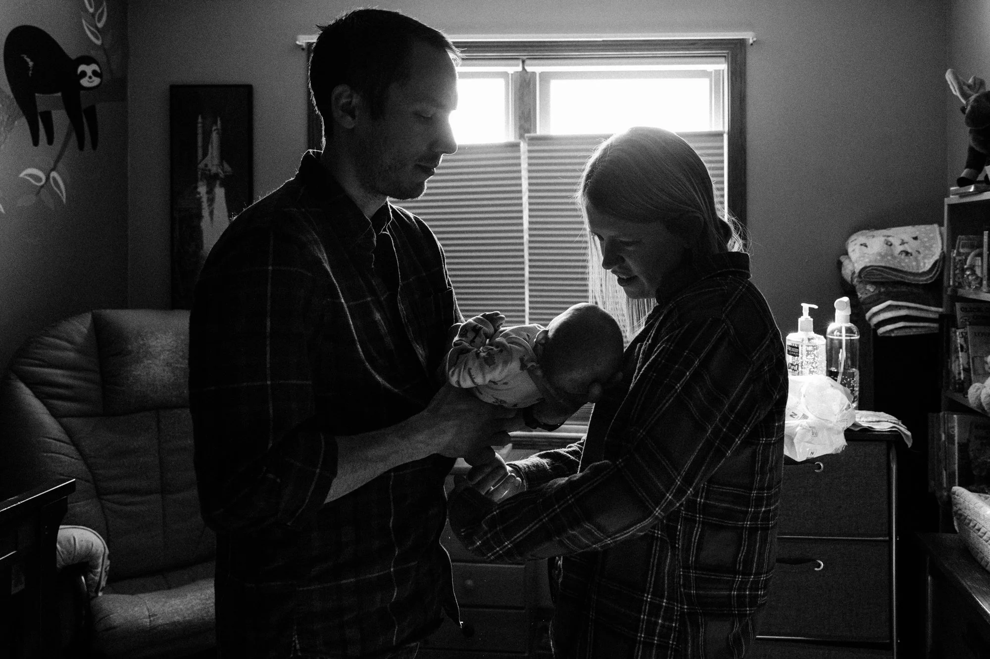 A couple holding their newborn baby in a bedroom with a window, shelves, and bottles of sanitizer in the background.