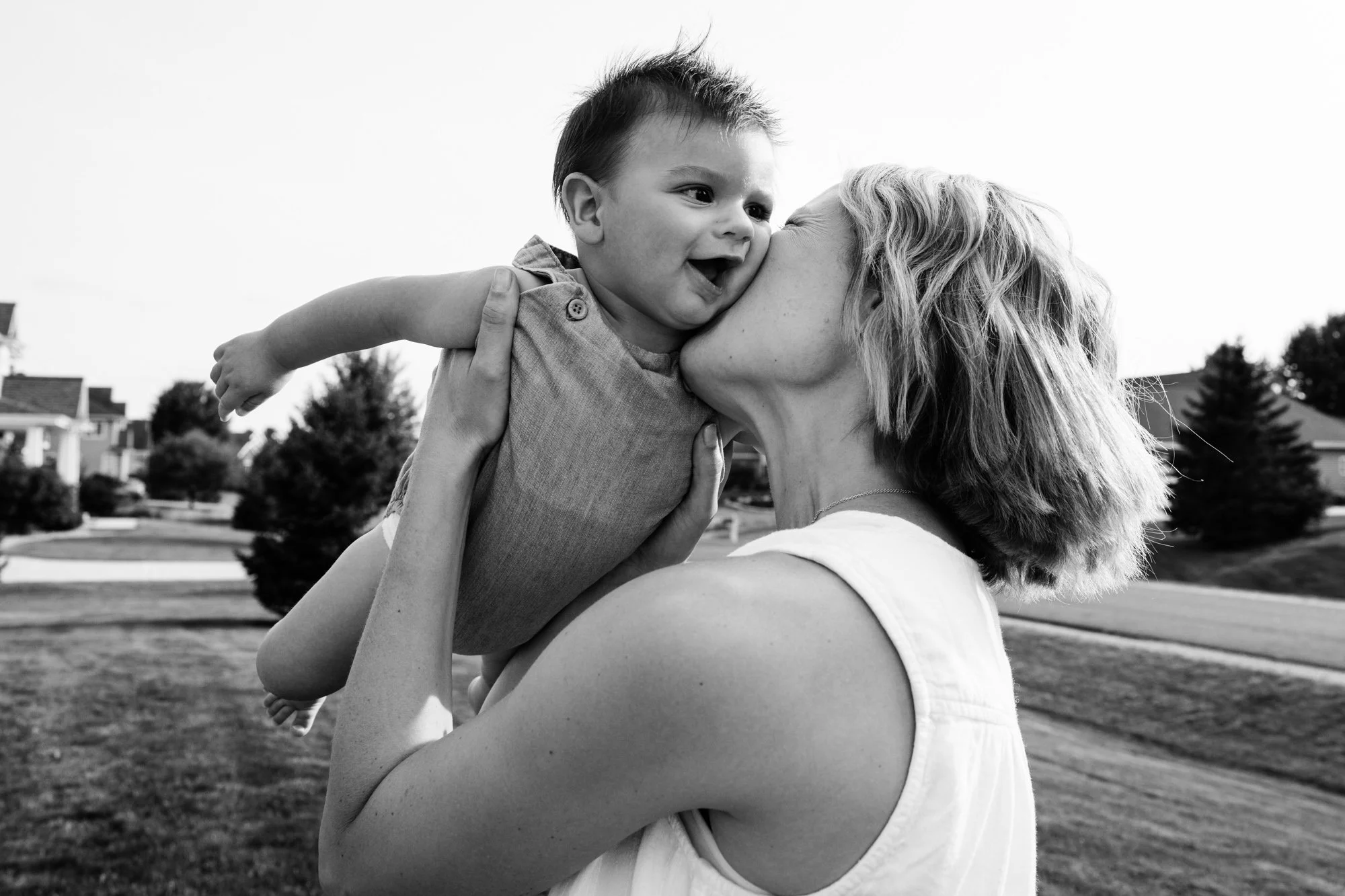 A woman with short hair holding a young child up to her face outside in a suburban neighborhood, both smiling.