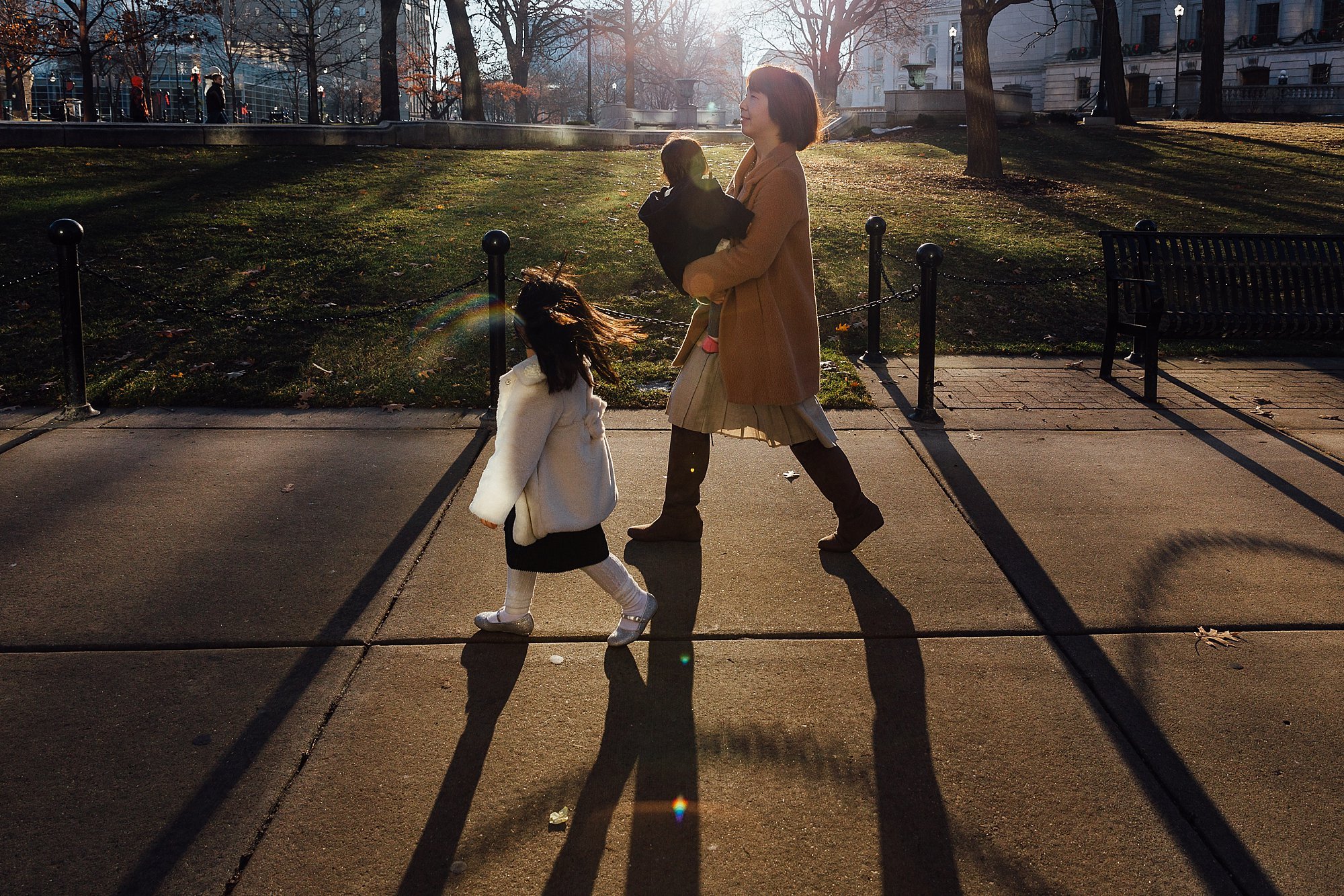 A woman walking with two young girls on a city sidewalk in the late afternoon, with sunlight casting long shadows. The woman is holding one girl and the other girl is walking ahead. Trees and buildings are visible in the background.