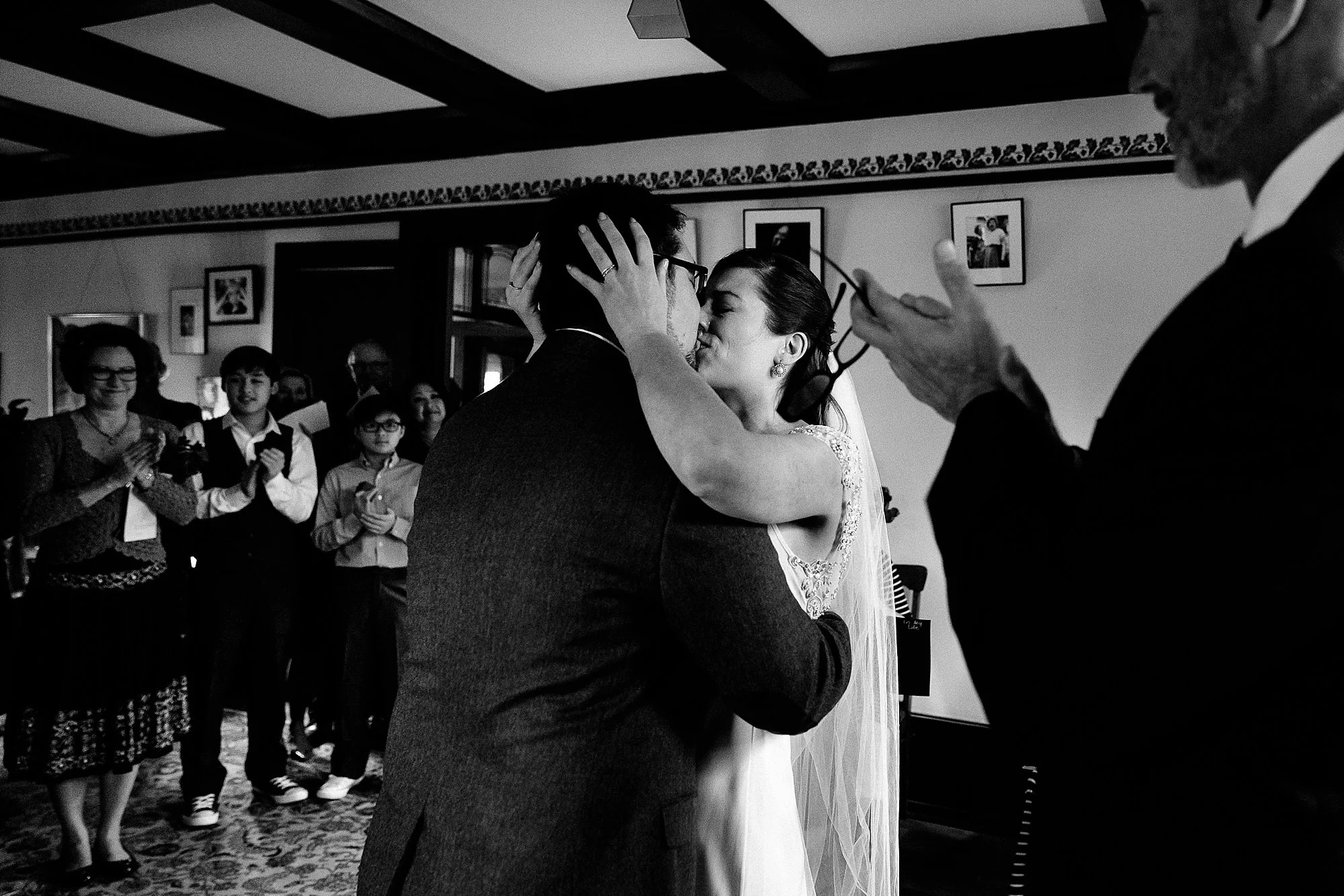 A black and white photo of a wedding ceremony where a couple is kissing. The bride, wearing a wedding dress and veil, has her hands on the groom's face. The groom, in a dark suit, has his arms around the bride. Several guests are in the background, c