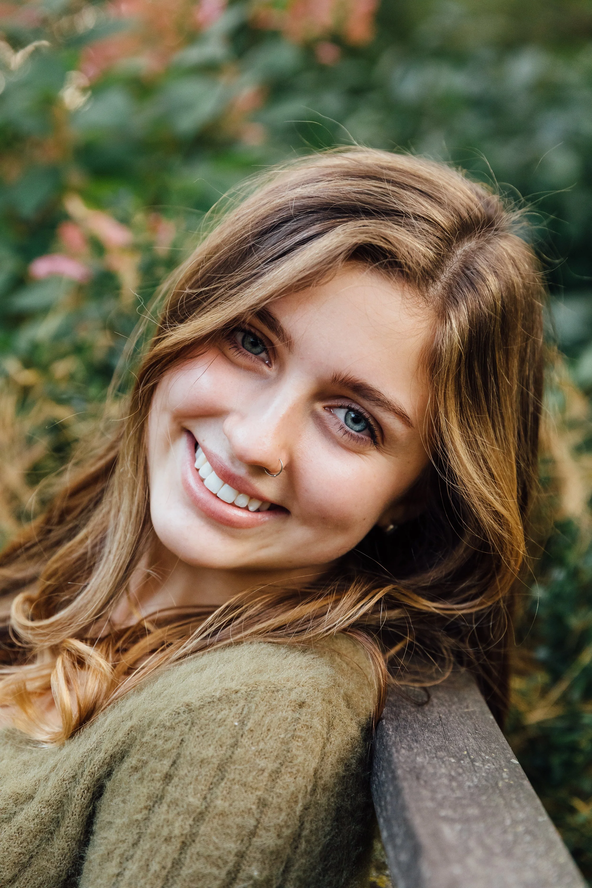 A smiling young woman with blue eyes and long, wavy brown hair, leaning against a wooden railing outdoors, surrounded by greenery.