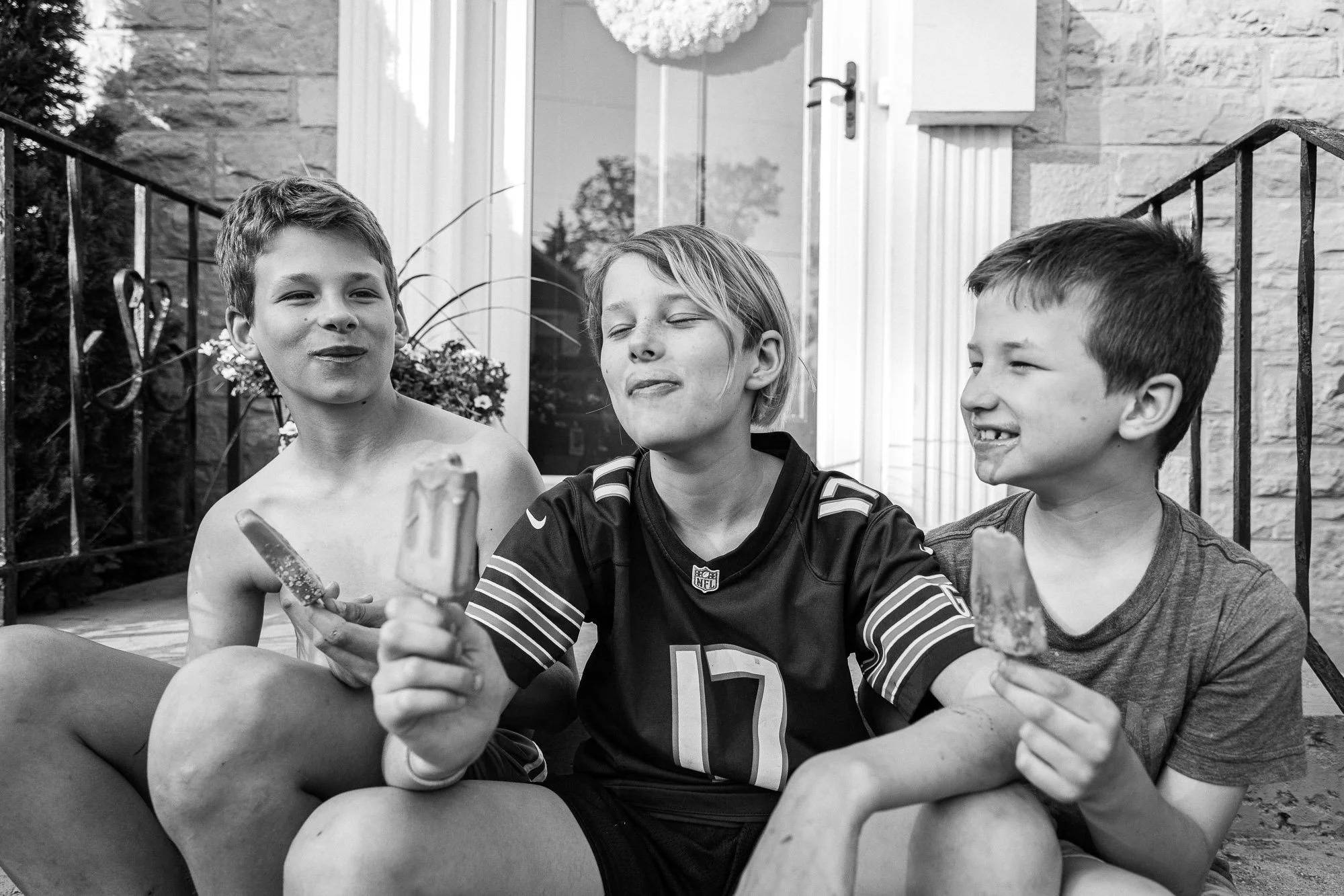 Three boys sitting outside on a porch eating ice cream cones, smiling and enjoying each other's company.