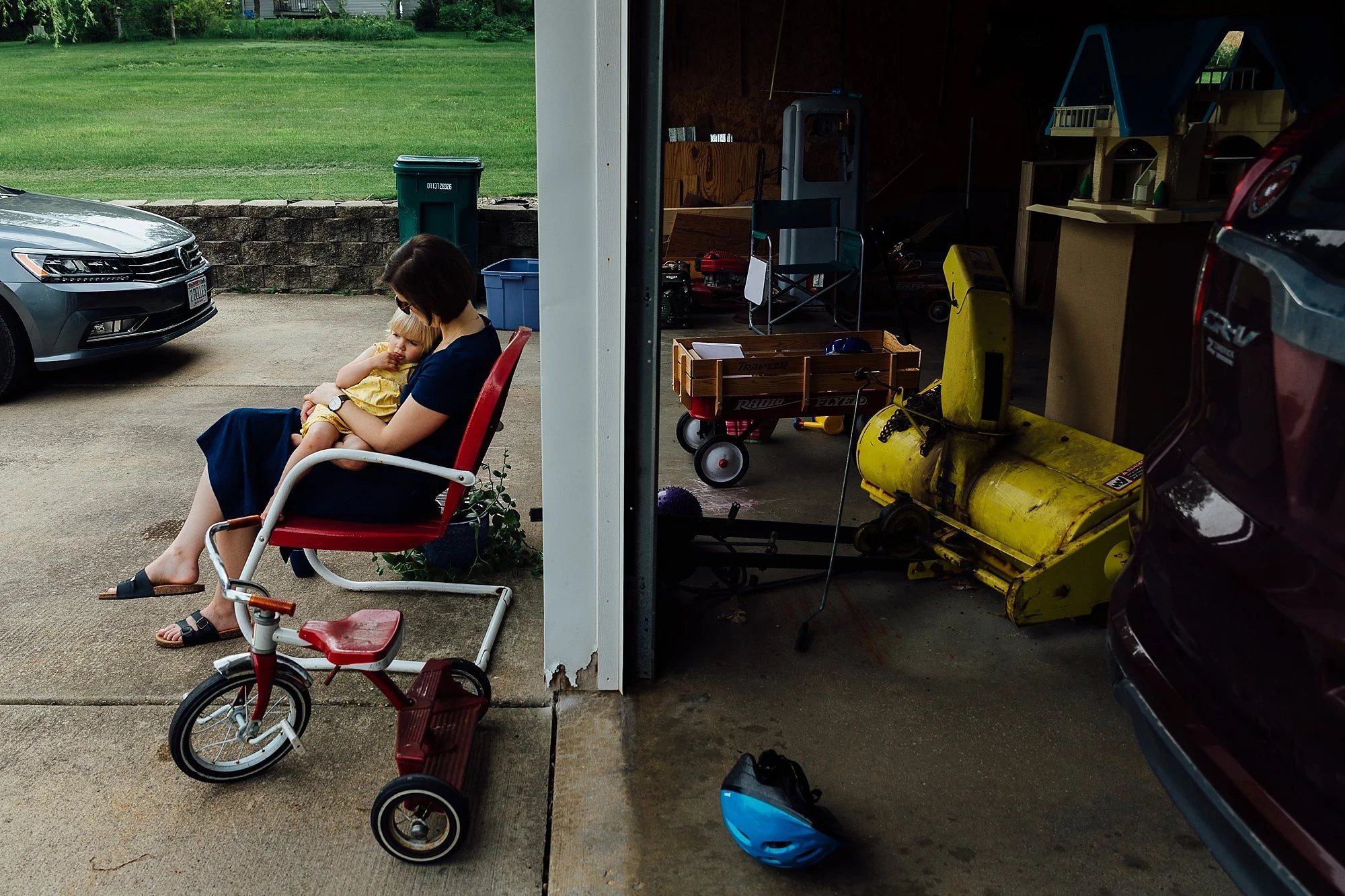 A woman sitting outside a garage on a red chair, holding a small child in her lap, with a tricycle nearby. The garage contains various tools and equipment, and there are parked cars and a grassy yard in the background.