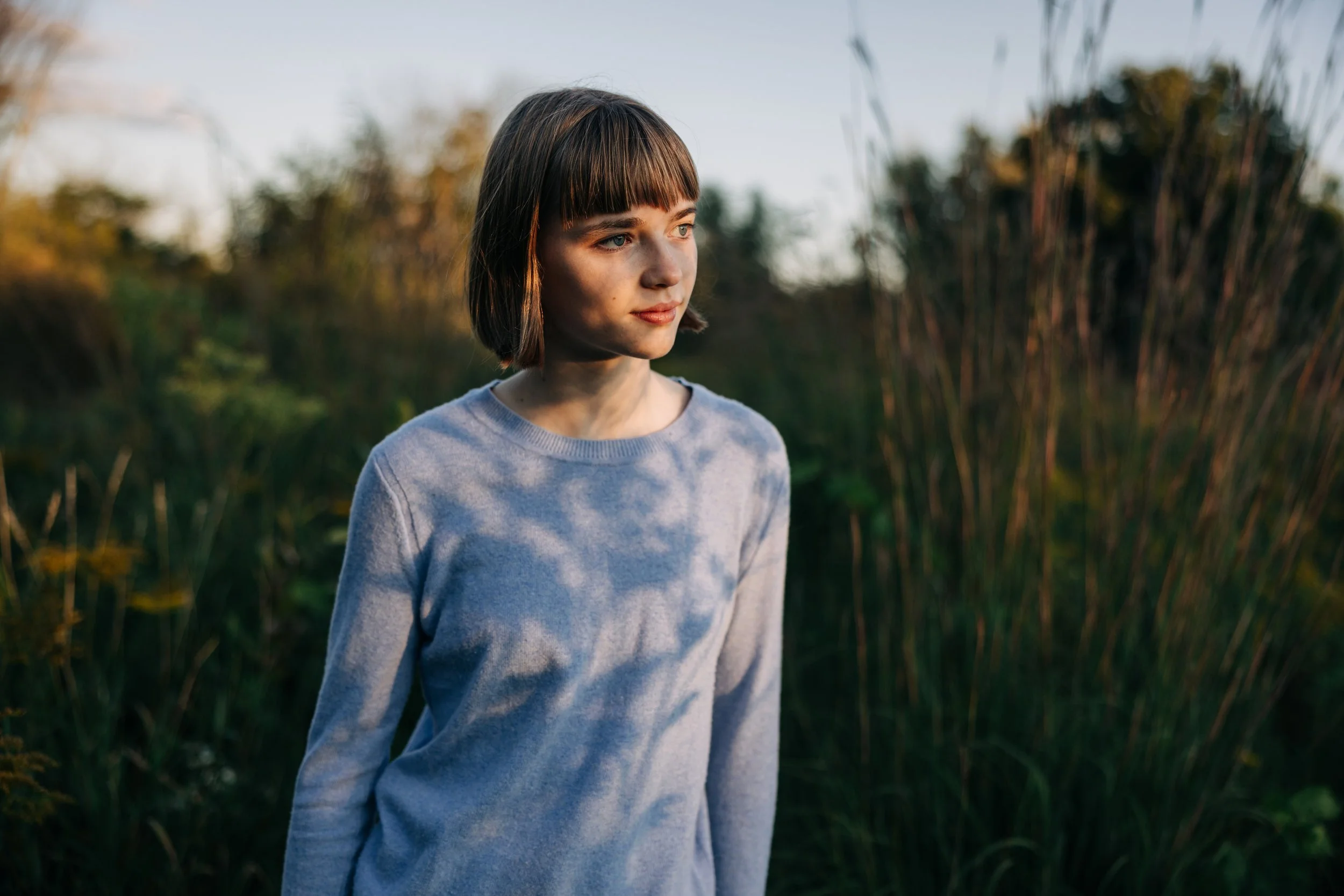 Young woman with a bob haircut wearing a gray sweatshirt standing outdoors in a natural setting during sunset.