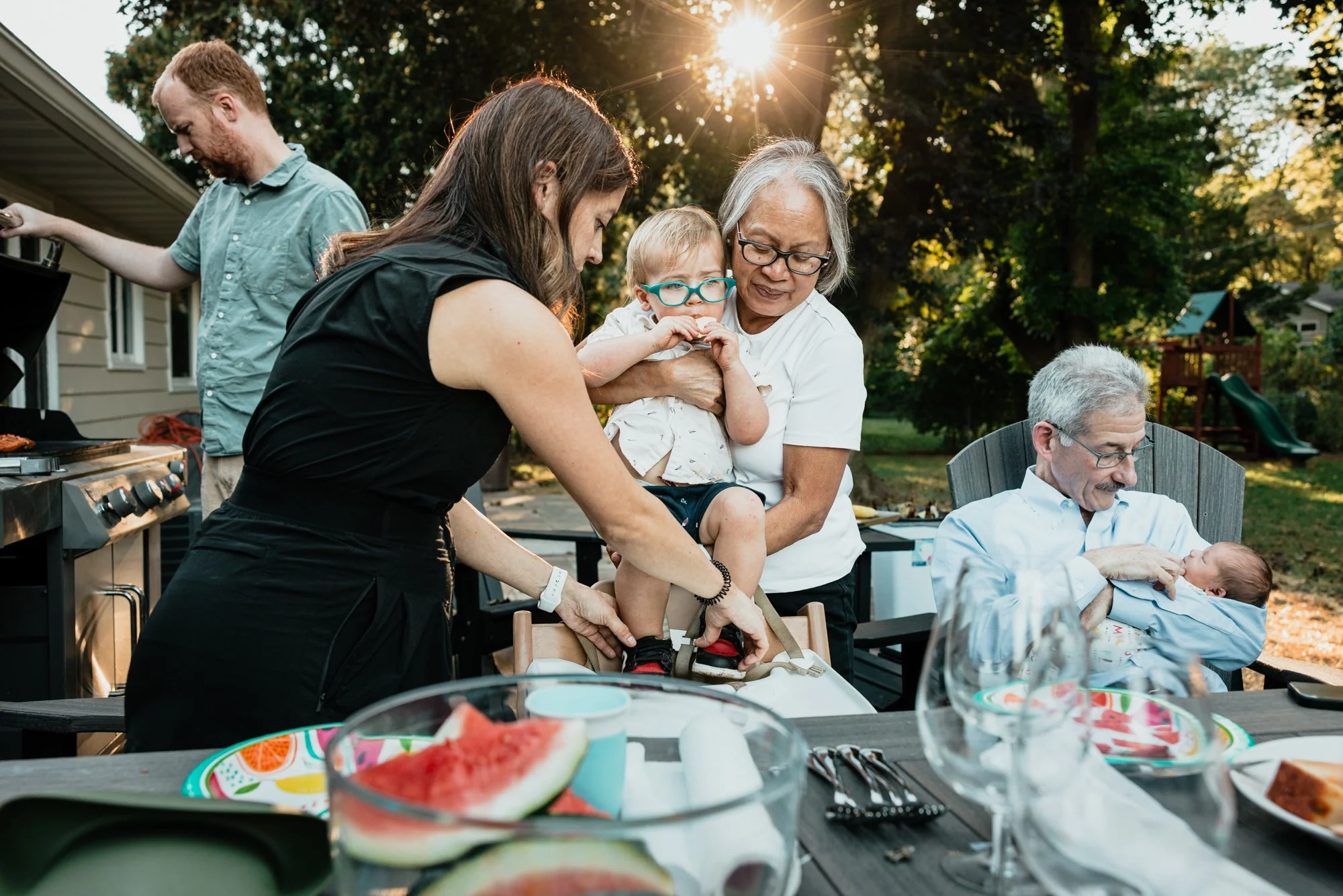 A family gathering around a dinner table outdoors in the evening, with a young woman helping a little boy put on a harness, an older woman holding a toddler, and an older man holding a newborn baby.