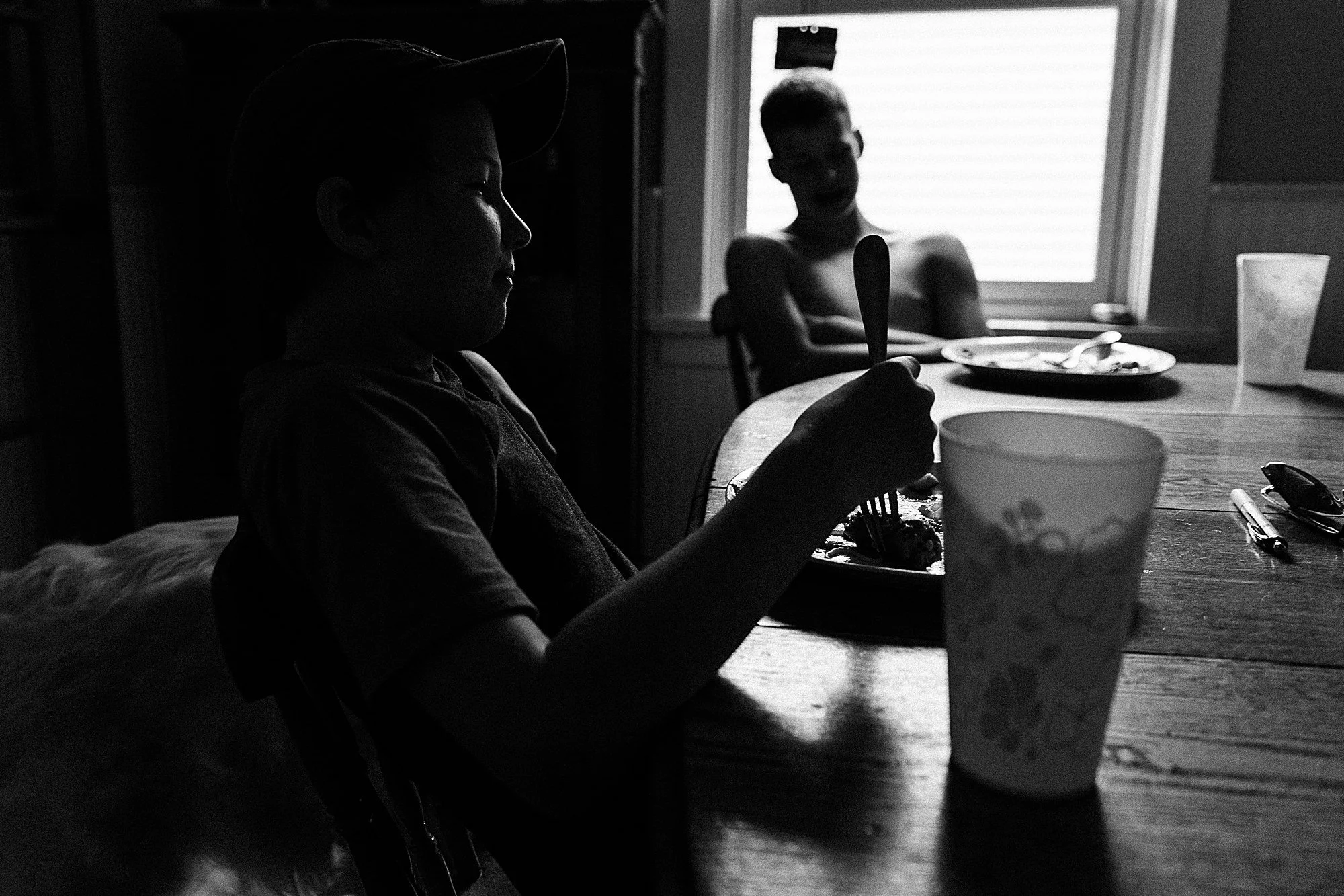 Two children sitting at a dining table, one in the foreground eating from a plate using a fork, with a cup in front, and one in the background smiling near a window.