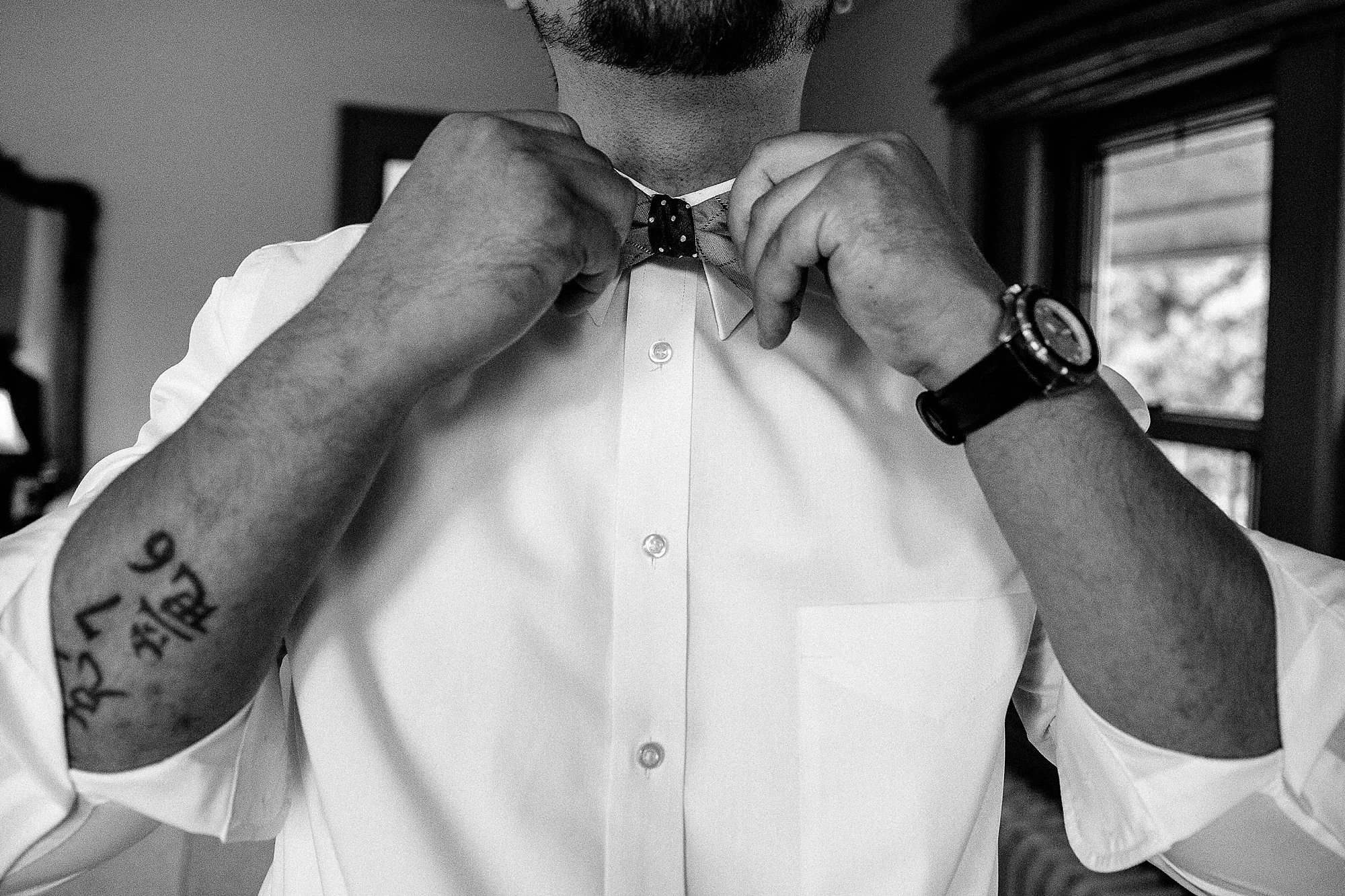 Man adjusting a bow tie, wearing a white shirt and a watch, with a tattoo on his arm, in a room with a window in the background.