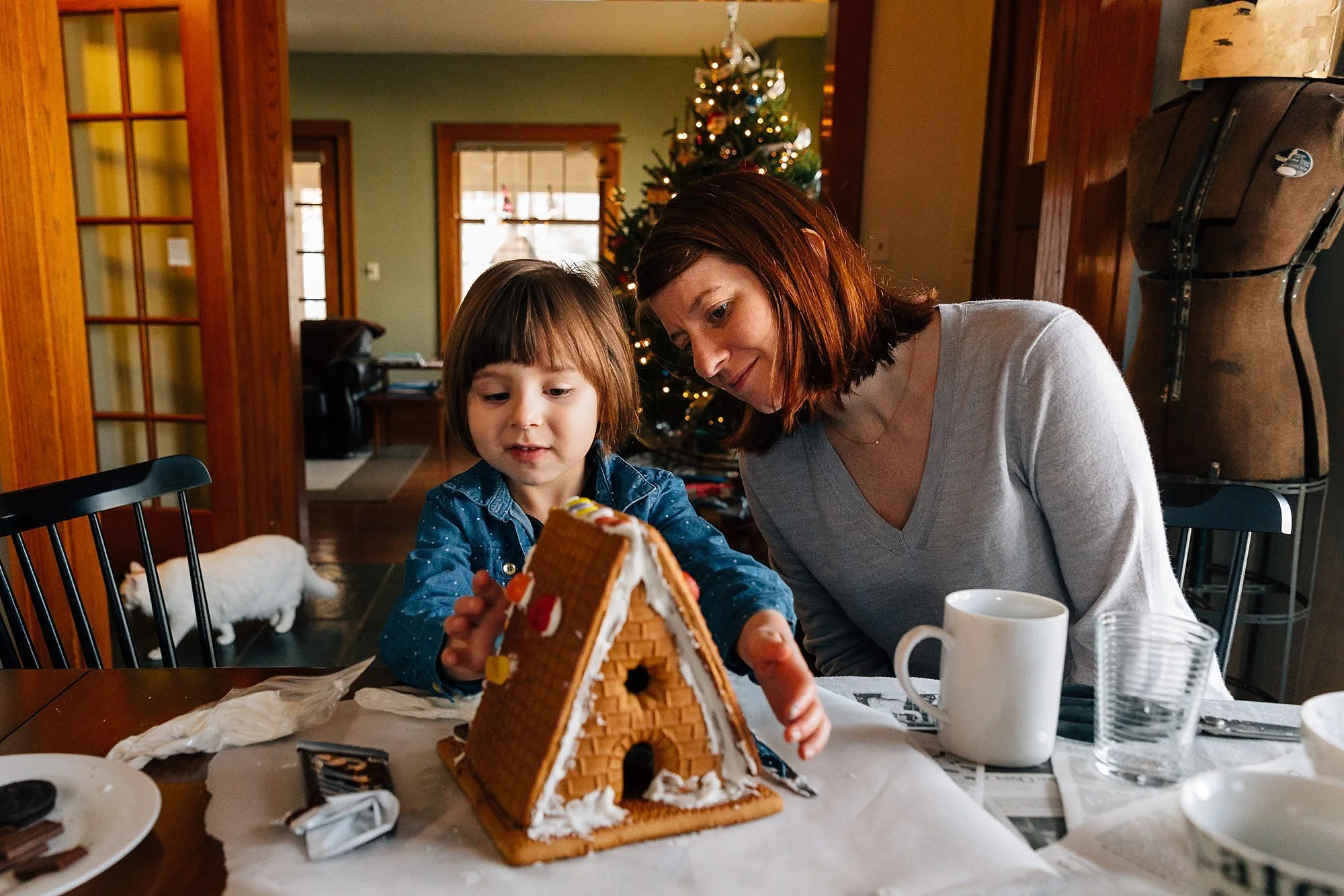 A woman and a young girl decorating a gingerbread house at a table during Christmas, with a Christmas tree and a white cat in the background.