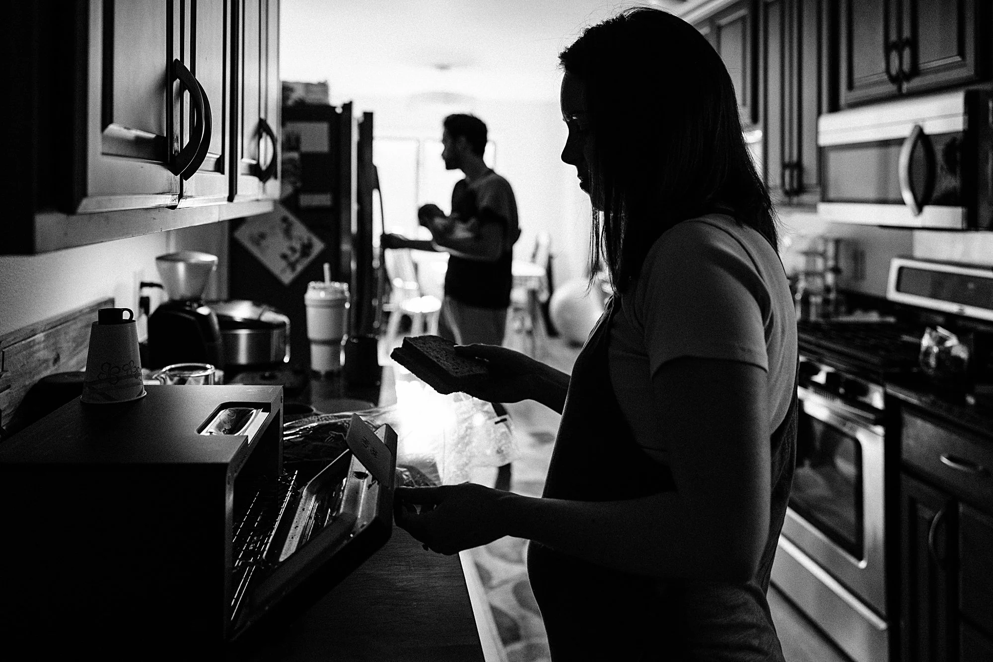 Silhouetted woman in kitchen holding toast, with two people in background, silhouetted against bright light.