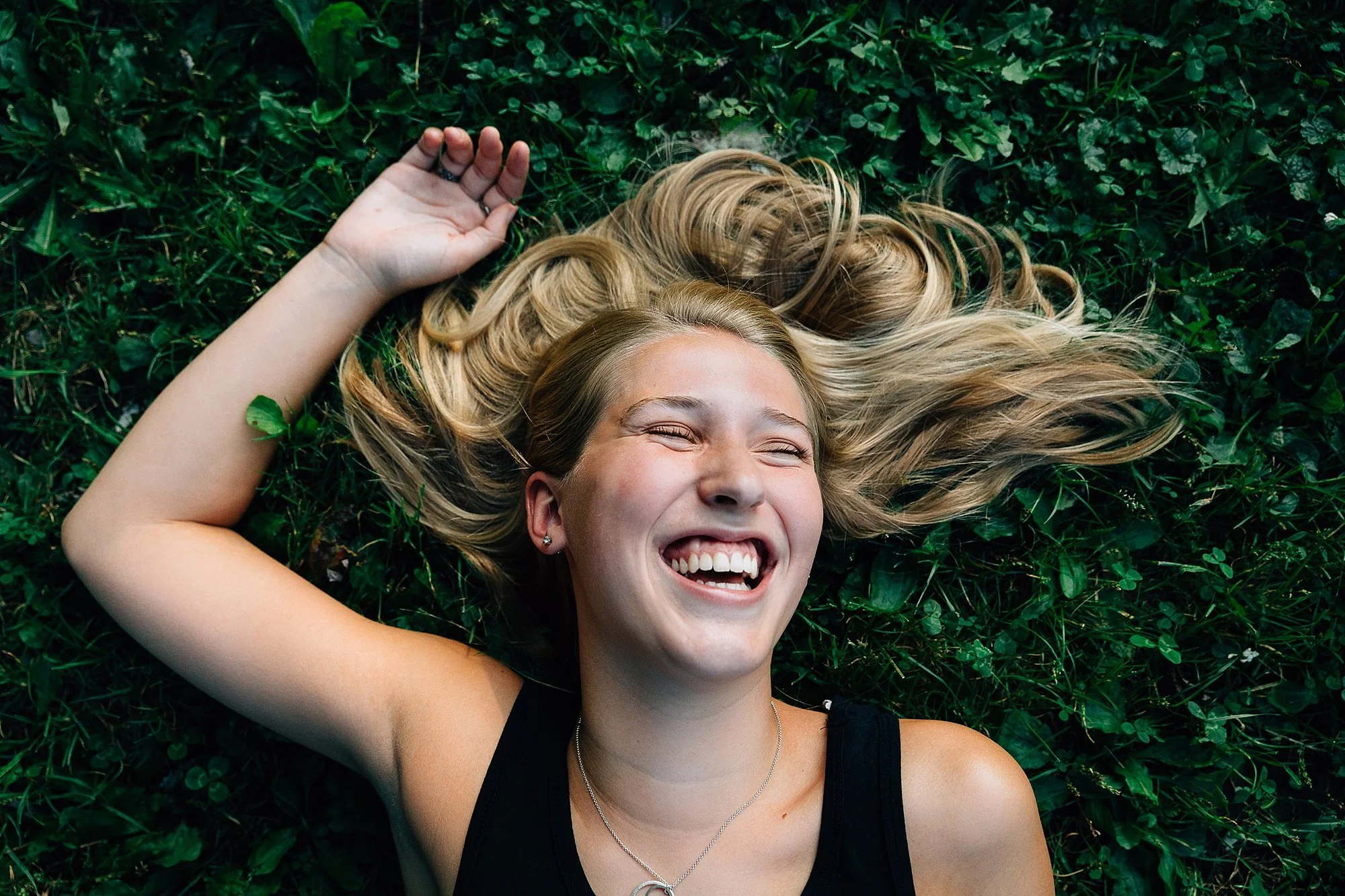 A young woman with blonde hair lying on green grass, smiling with eyes closed, wearing a black tank top and a necklace.