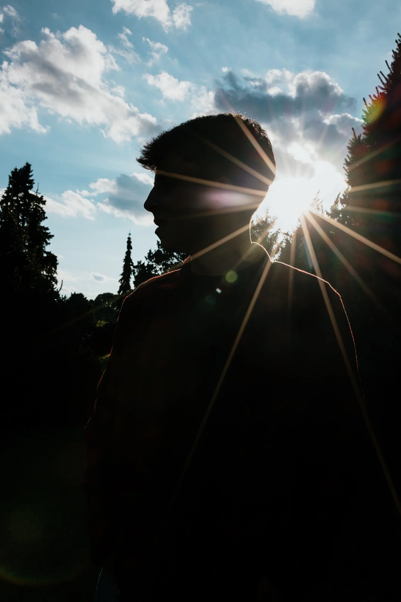 Silhouette of a person standing outdoors with trees and cloudy sky behind, sunlight creating a starburst effect around the person's head.