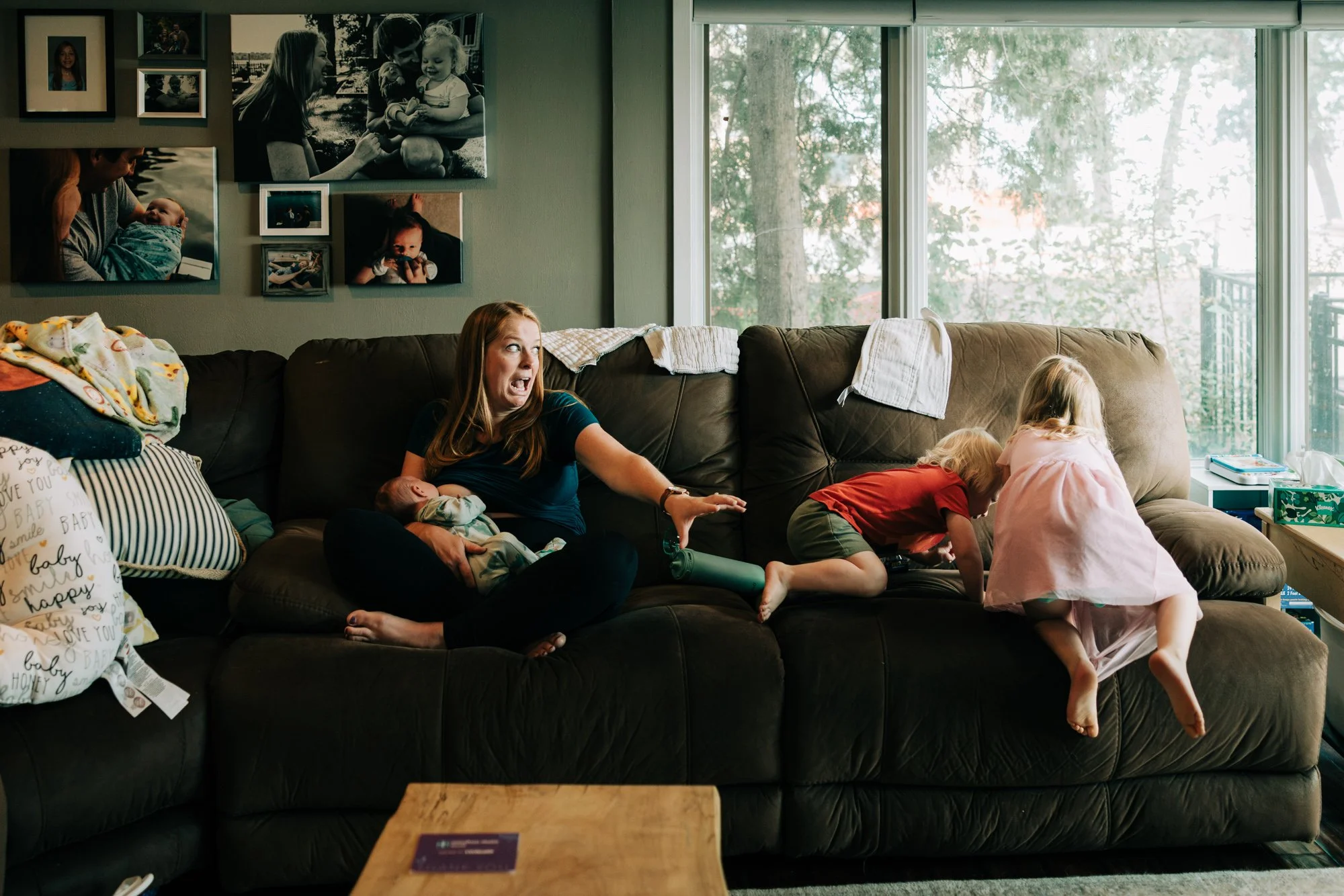 Mother sitting on a brown sofa holding a newborn baby, with two young children climbing on the sofa beside her, in a living room.