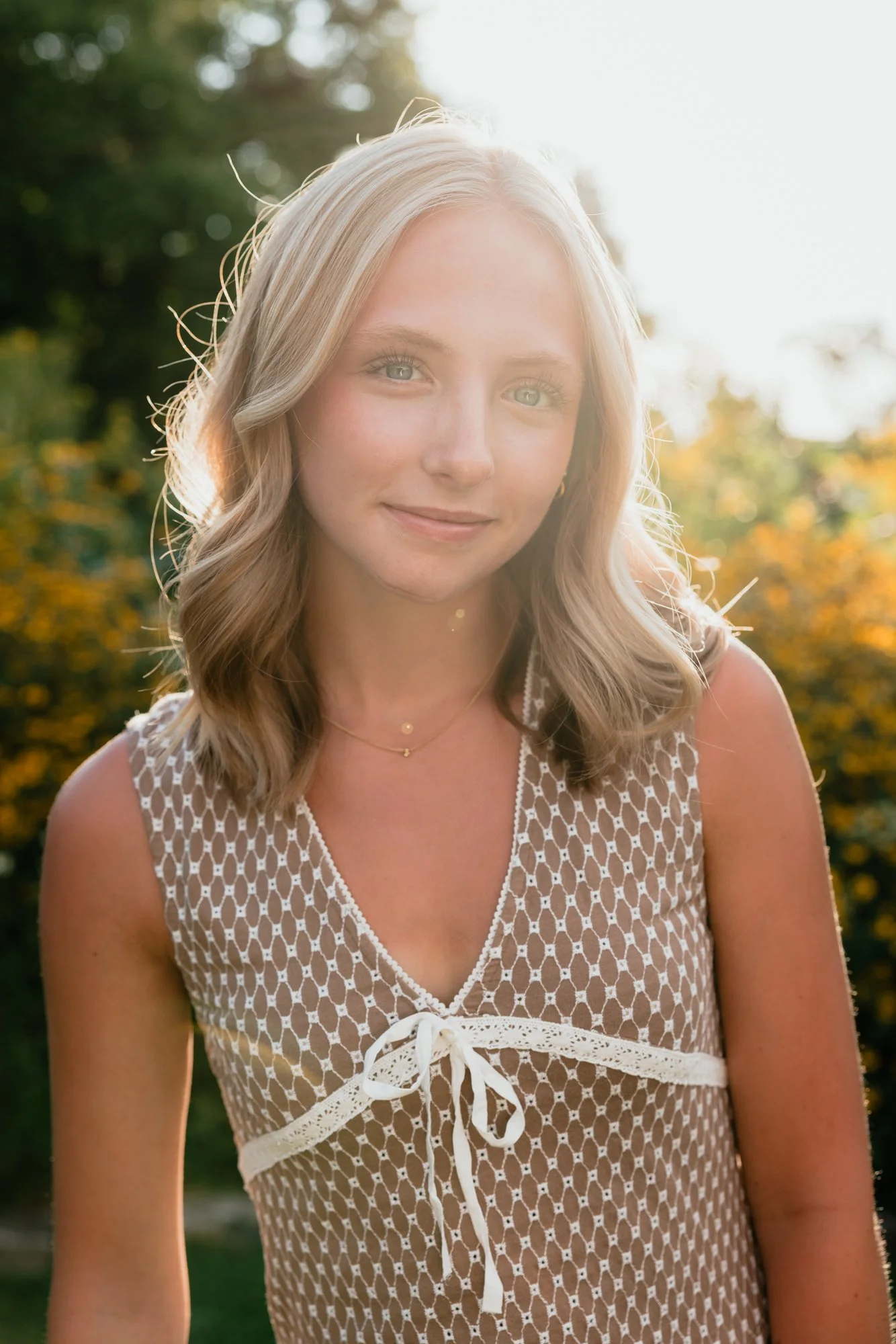 A blonde woman with wavy hair, wearing a sleeveless white lace dress with a bow tie at the front, smiling outdoors with sunlight shining behind her, and trees in the background.