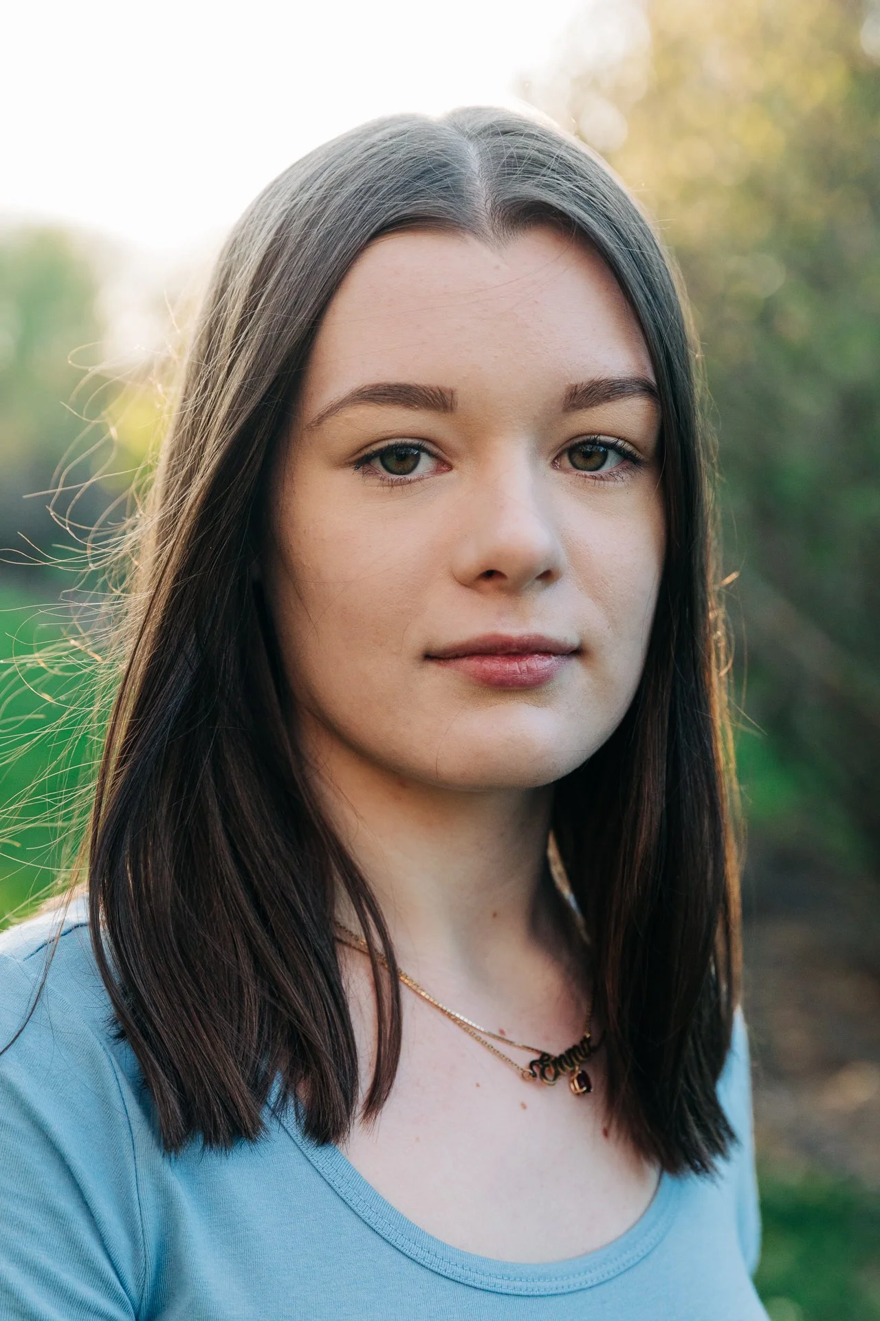 A young woman with shoulder-length dark brown hair, wearing a light blue shirt and a gold necklace with the name 'Emma,' stands outdoors during daylight with a blurred background of green trees and sunlight.
