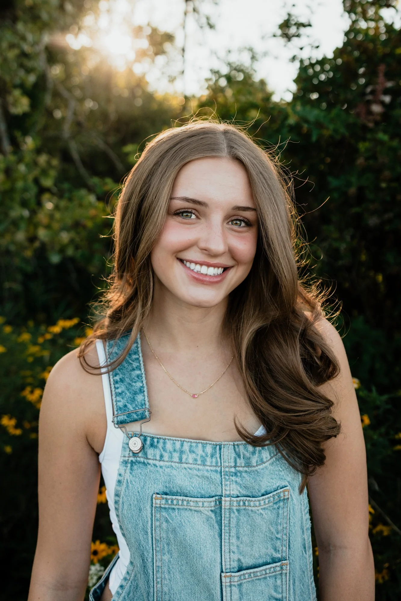 Young woman with long brown hair, smiling outdoors during sunset, wearing denim overalls and a white top, with greenery and yellow flowers in the background.