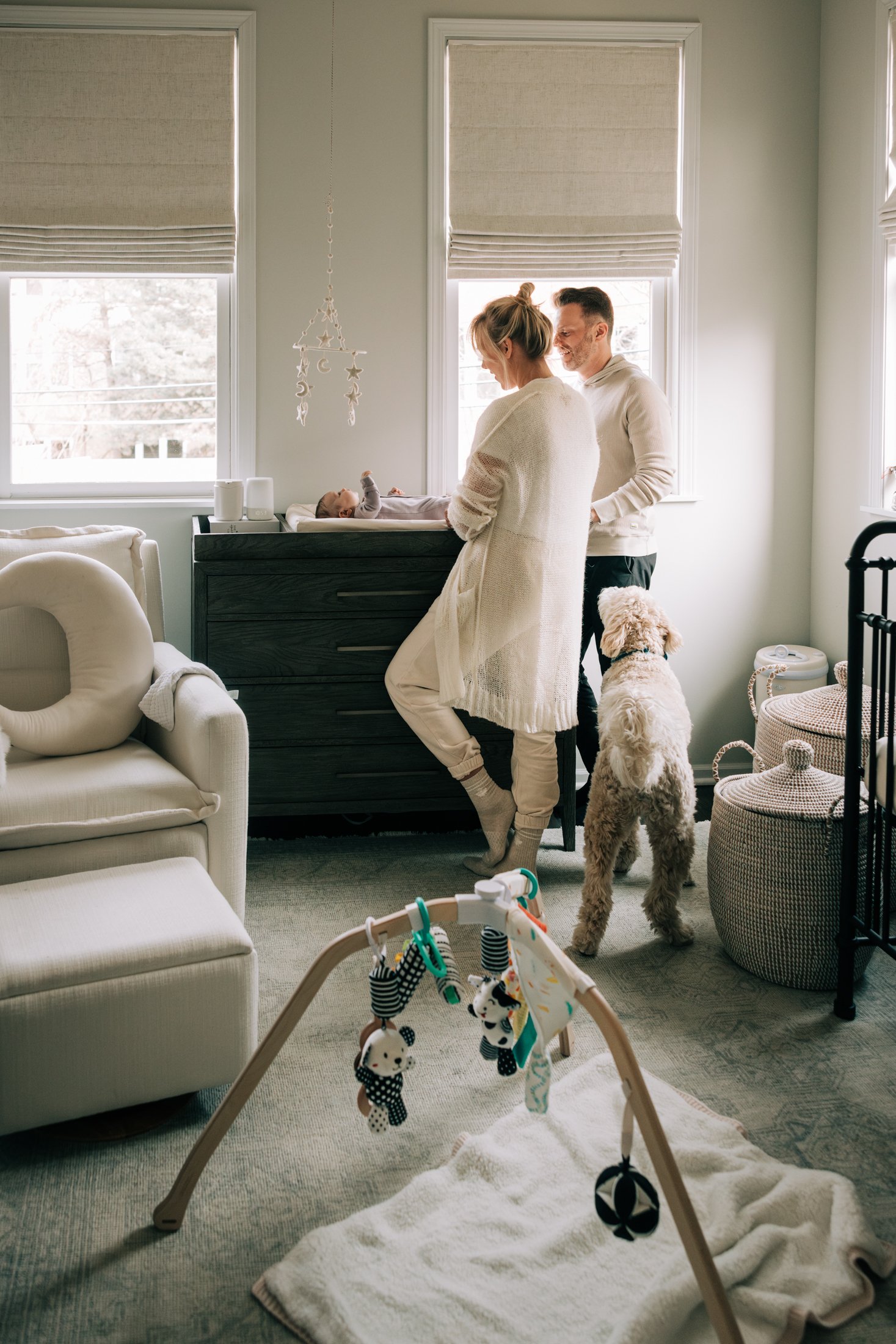 A couple with a dog watching their baby in a crib in a cozy, light-colored living room with large windows, a changing table, and playful baby toys.
