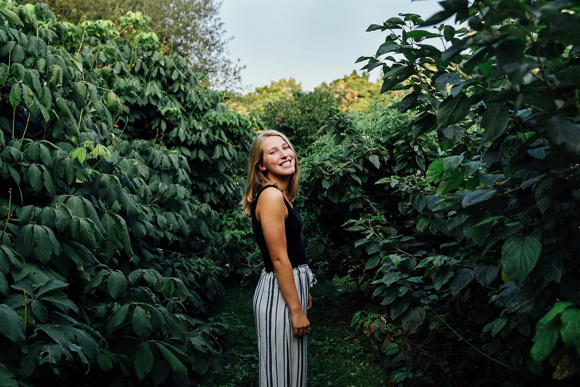 A young woman with blonde hair smiling, standing in a lush green garden path surrounded by green foliage and plants.