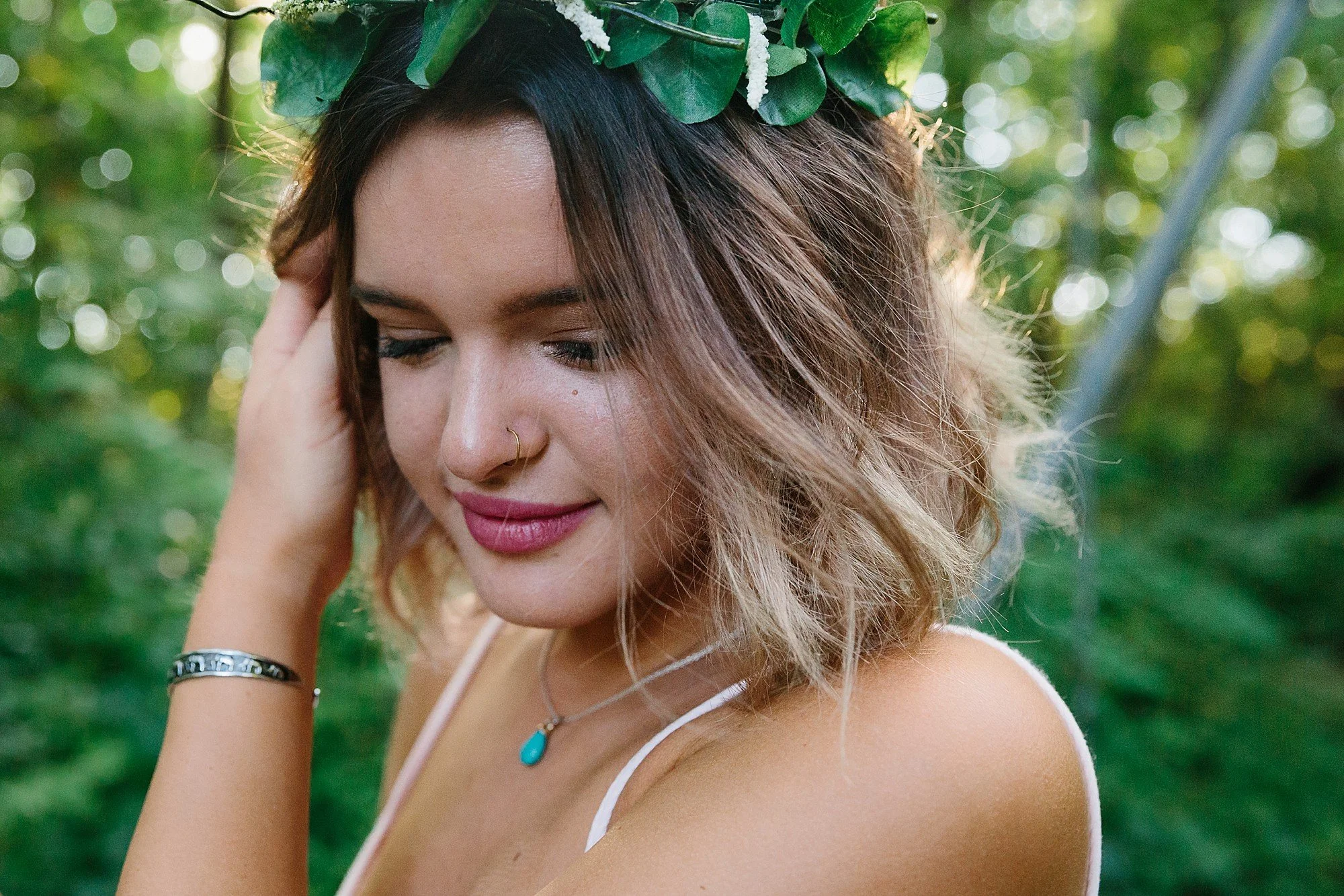A young woman with shoulder-length, wavy hair, floral headpiece, and a nose ring, smiling with eyes closed outdoors surrounded by greenery.