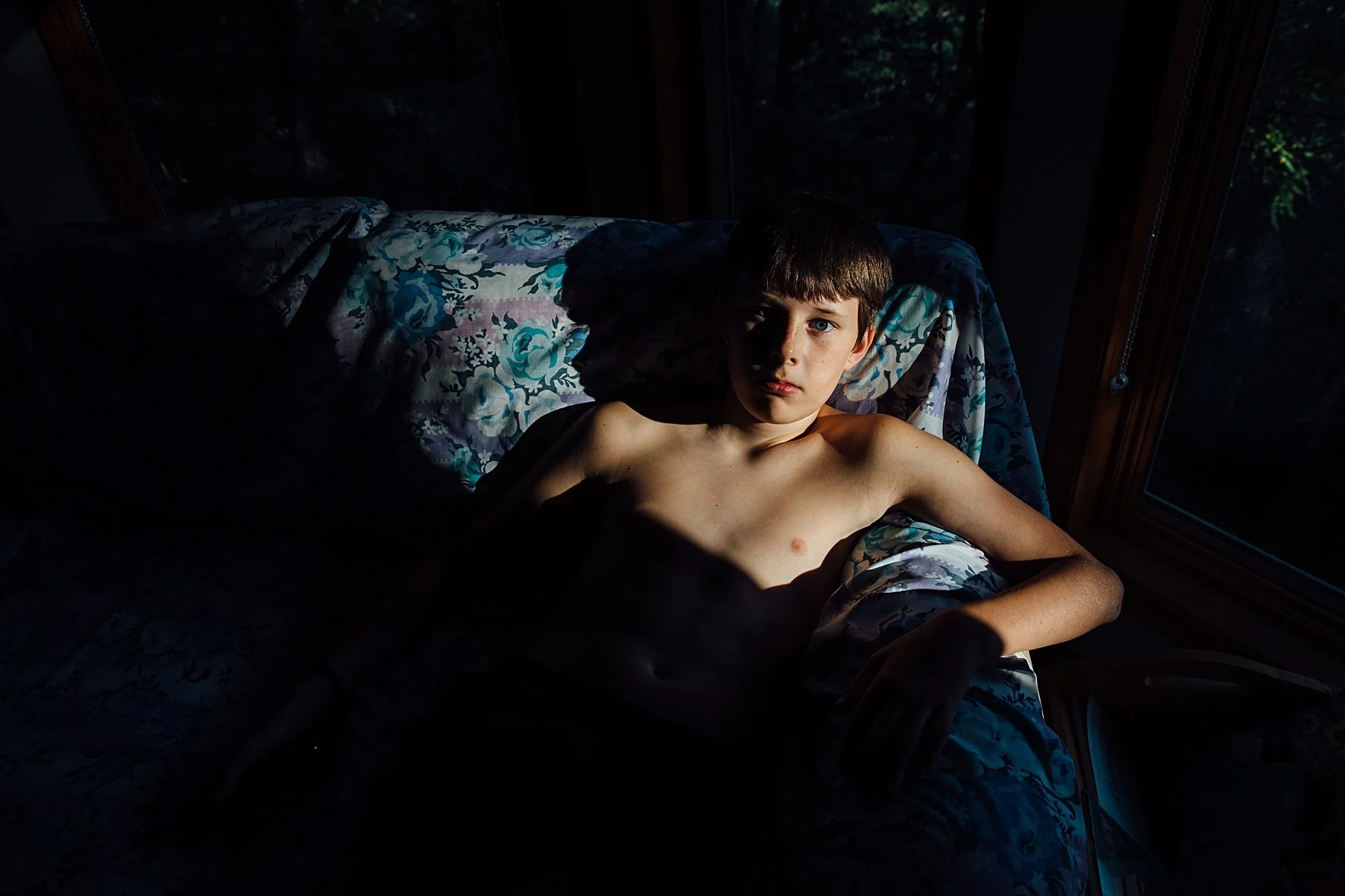 A young shirtless boy with brown hair and blue eyes lying on a bed, illuminated partially by natural light coming through a window, with dark shadows on part of his face and body.
