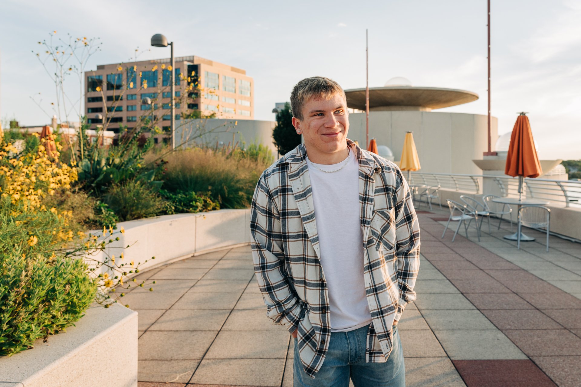 Young man standing outdoors on a rooftop terrace during sunset, smiling with his hands in his pockets, wearing a plaid shirt over a white t-shirt and jeans, with plants, chairs, and buildings in the background.