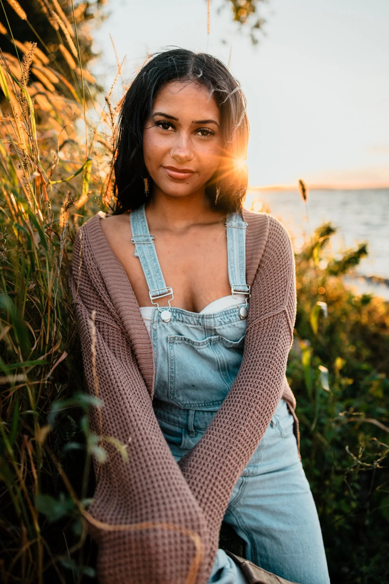 A woman with dark hair wearing a beige knit sweater and denim overalls sitting outdoors during sunset by a body of water with tall grass around her.