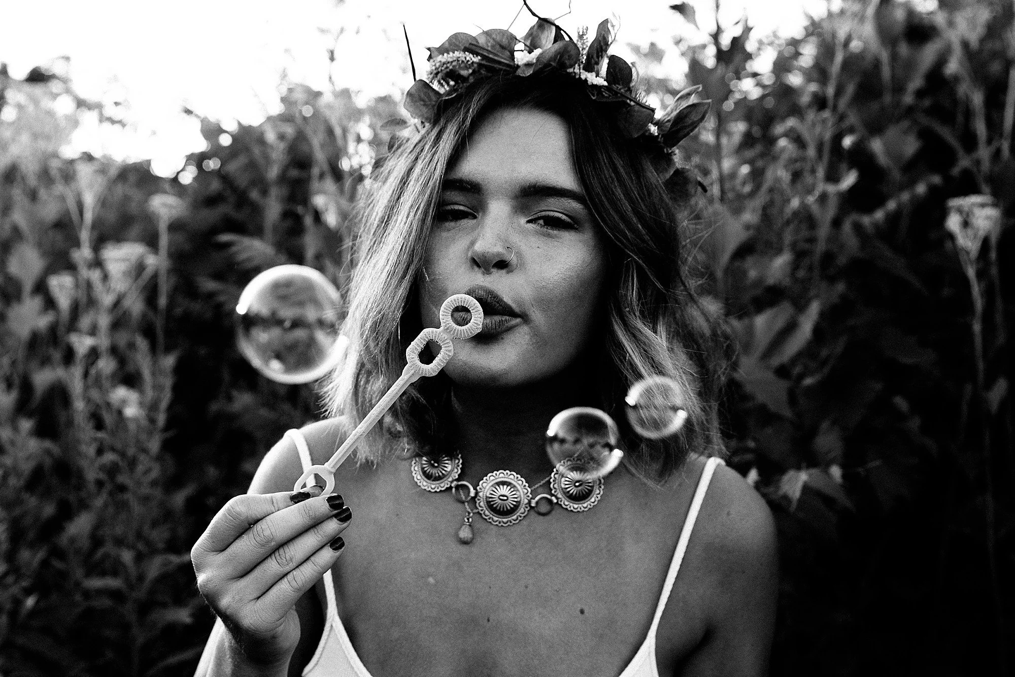 A woman with a flower crown blowing bubbles outdoors in a natural setting, captured in black and white.